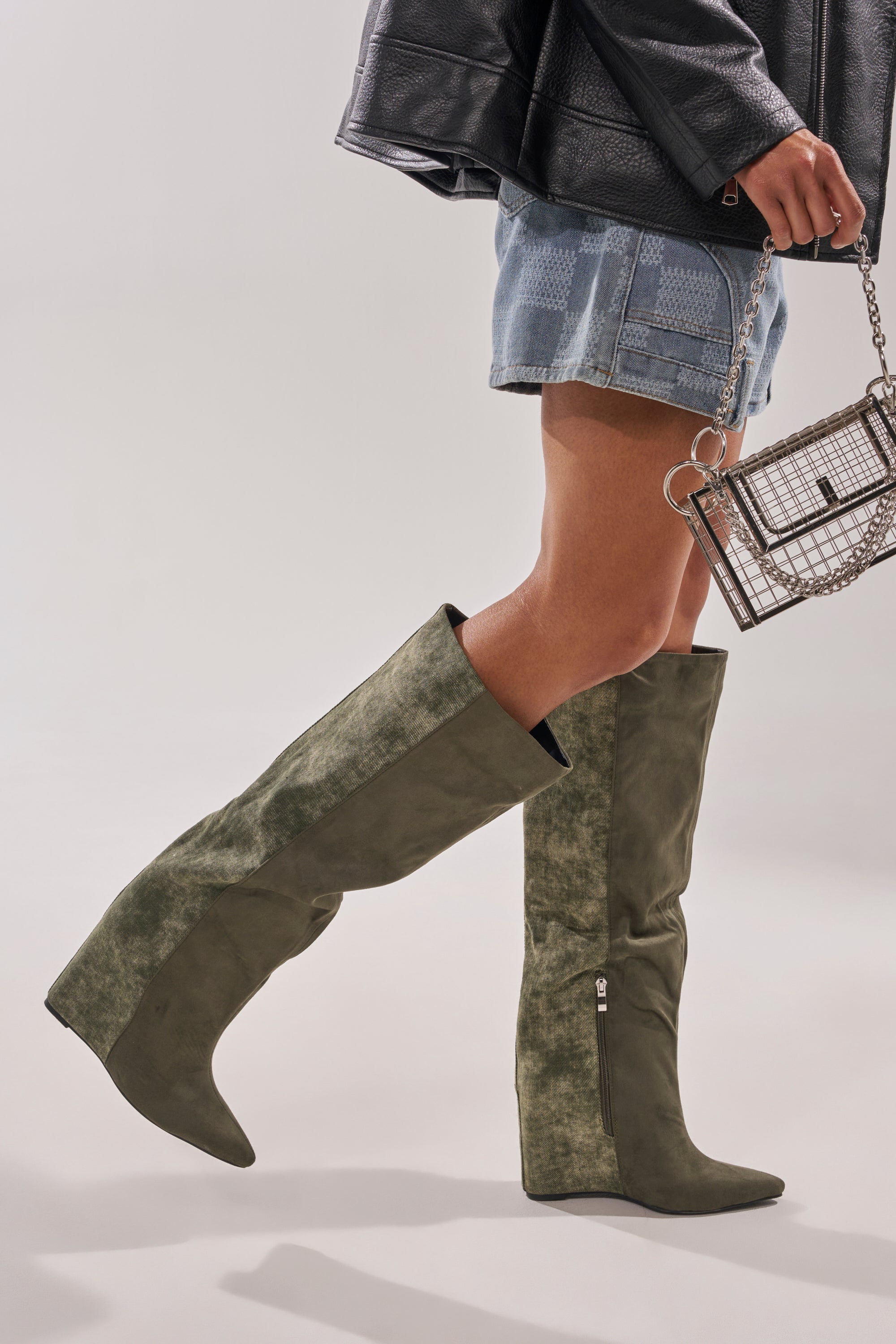 Wearing the AZALEA WANG GIANNA OLIVE TWO TONED BOOT, denim shorts, and a black leather jacket, a person holds a silver mesh handbag against a plain light background. 1