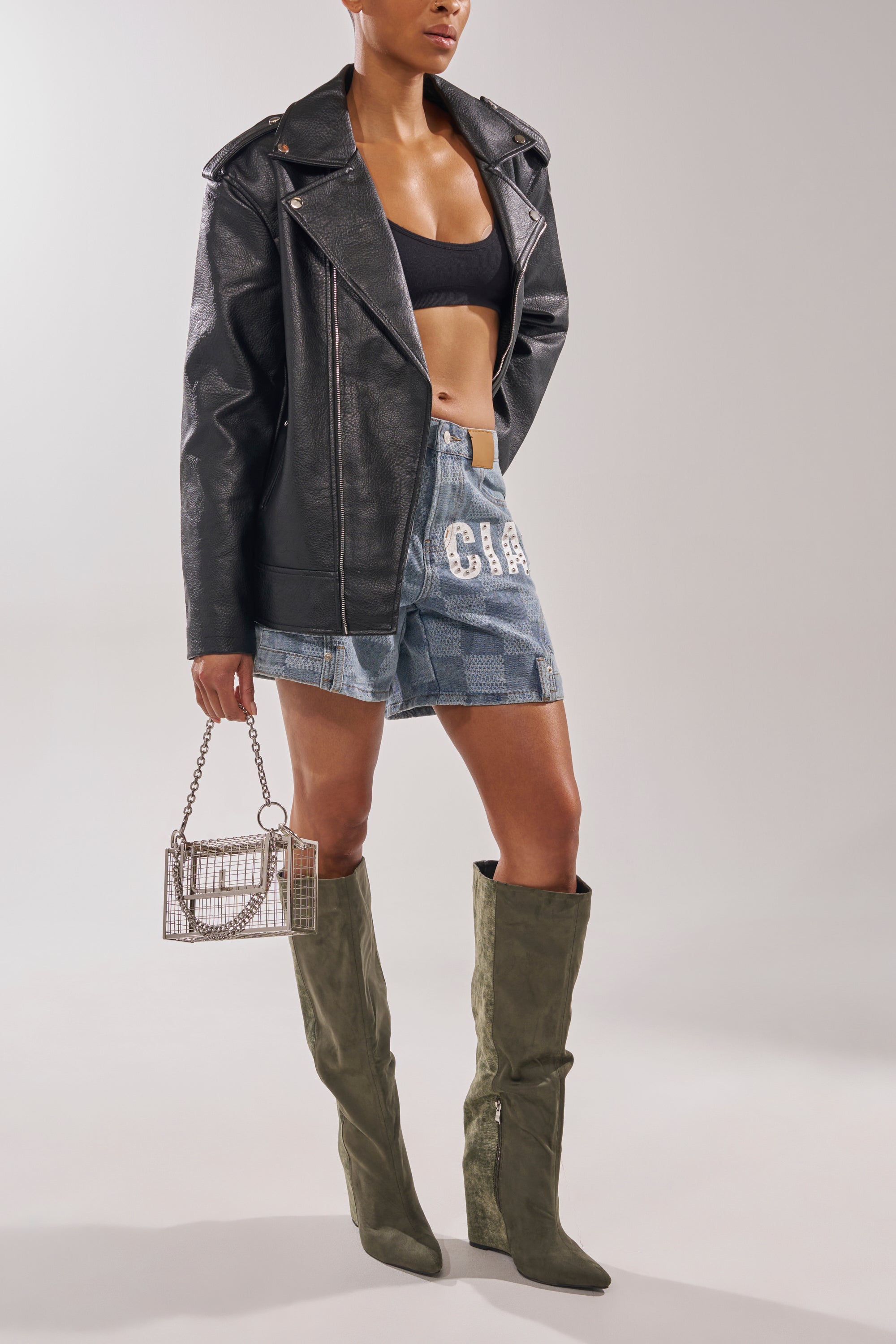 A person wearing a black crop top, oversized black leather jacket, denim shorts with white embellishments, holding a small metallic handbag, and rocking the AZALEA WANG GIANNA OLIVE TWO TONED BOOT. 2