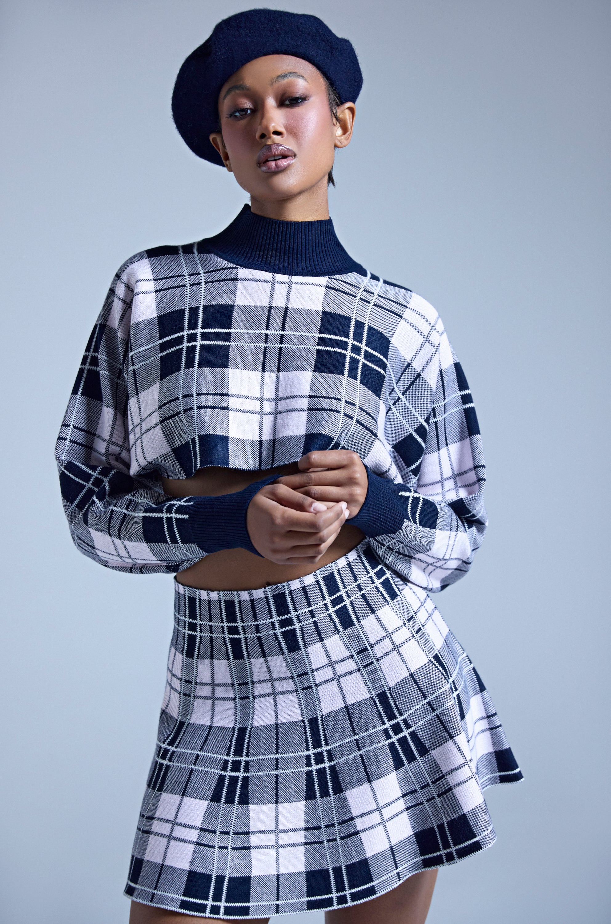 A person wearing the HOME FOR THE HOLIDAYS CROPPED SWEATER and a matching high-waisted plaid skirt poses confidently against a light blue background. 0