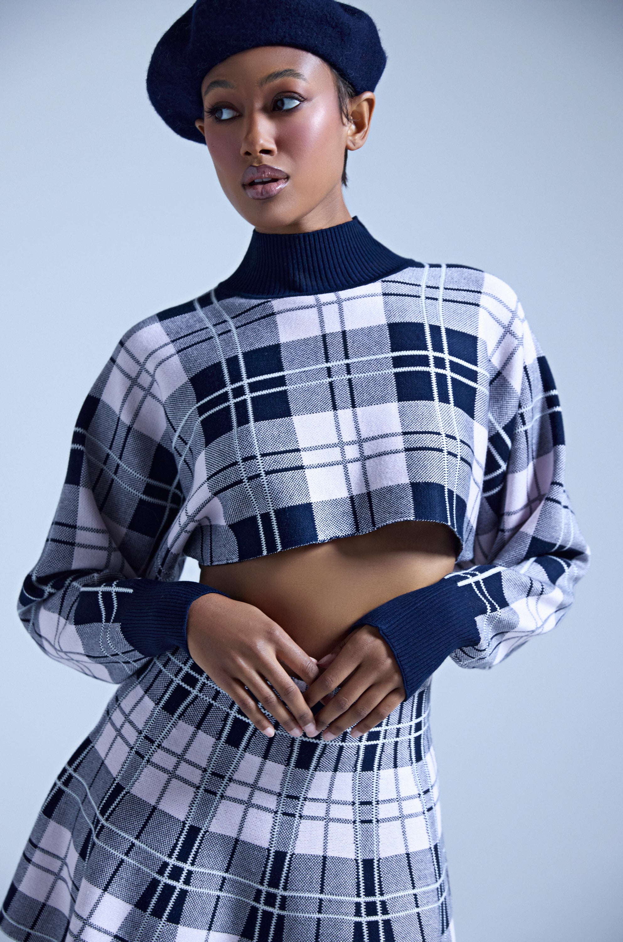 A woman poses with her hands near her waist against a light blue background, wearing the HOME FOR THE HOLIDAYS CROPPED SWEATER in navy blue paired with a matching plaid skirt and beret. 1