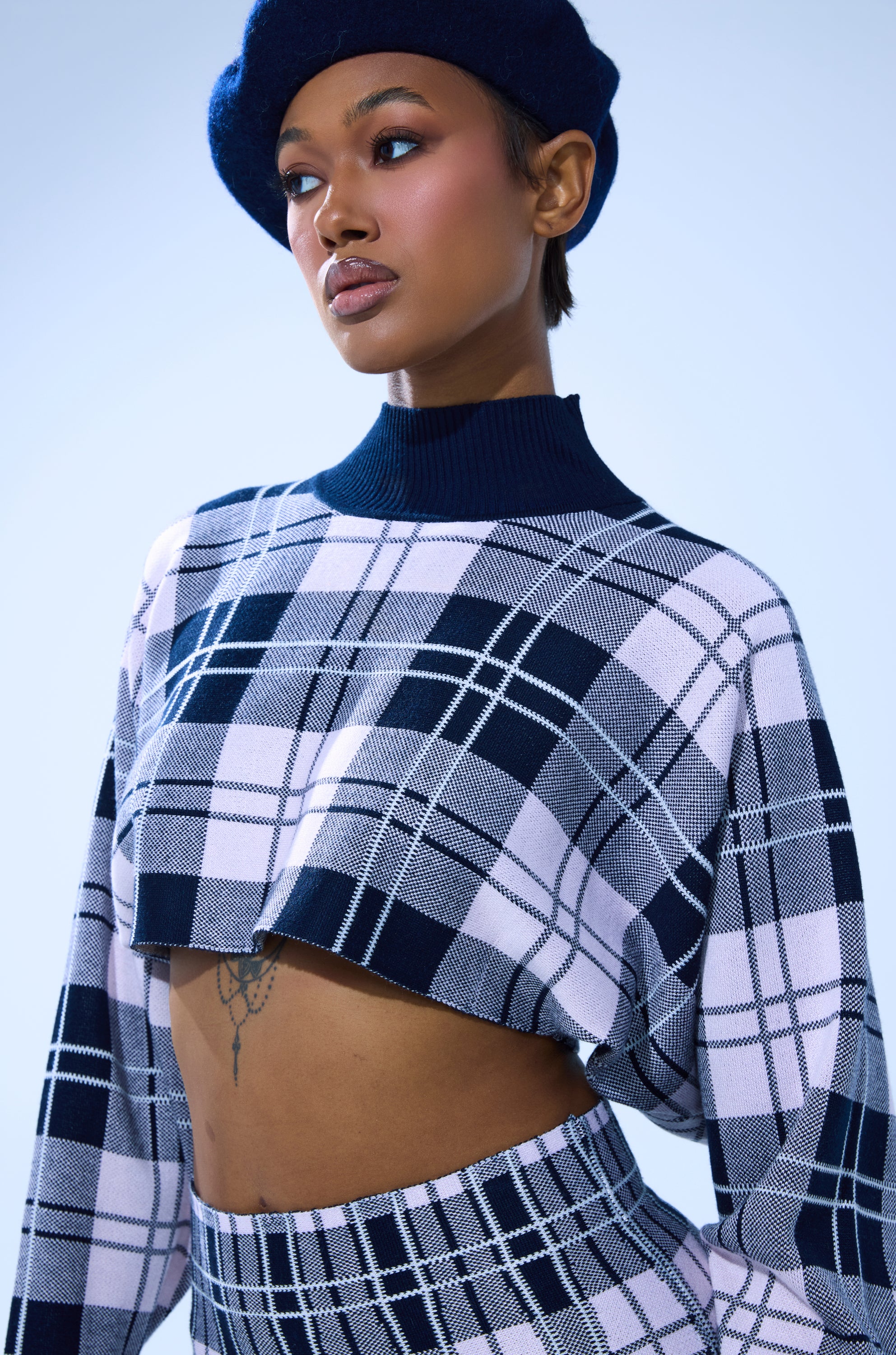 A woman in the HOME FOR THE HOLIDAYS CROPPED SWEATER and matching high-waisted pants stands against a light blue background, wearing a navy beret and looking confidently to the side. 2