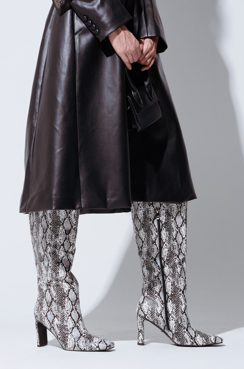 A person wearing a long black leather coat and the AZALEA WANG GILROY SNAKE CHUNKY HEEL BOOT stands against a light background, holding a small black handbag.