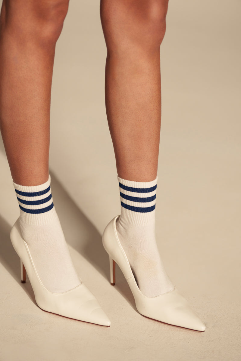 Wearing the AKIRA GINNY WHITE PUMP with white socks featuring blue stripes, a person stands on a light-colored floor; only legs and feet are visible. 0