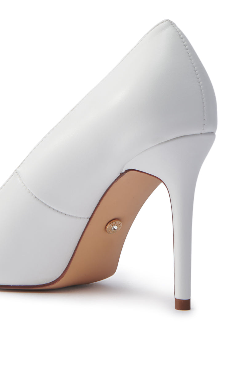 Close-up of the AKIRA GINNY WHITE PUMP highlighting its sleek stiletto heel and smooth faux leather upper. The focus is on the shoe's right side, showcasing a small metallic detail near the heel on the sole. 6