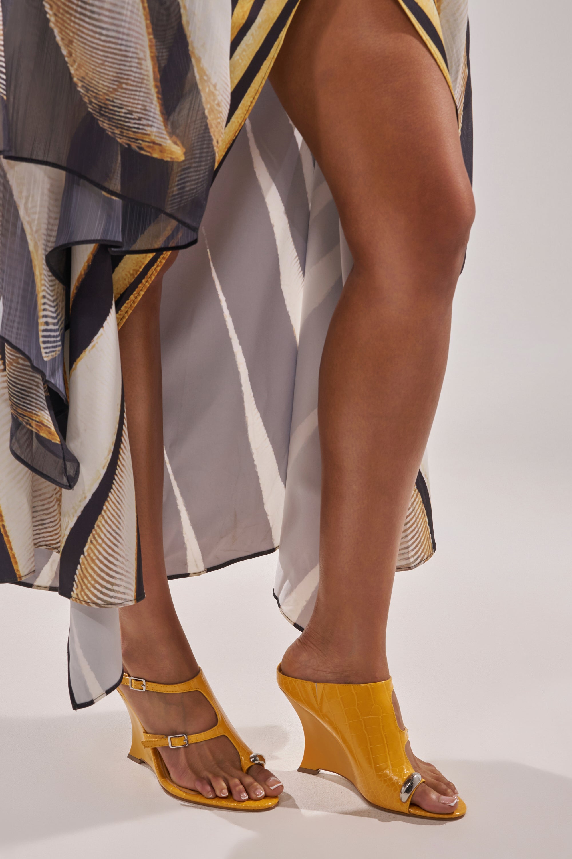 A person wears a flowing, patterned dress with yellow, black, and white stripes and AKIRA GOLD GINO MUSTARD CROC WEDGE SANDALS featuring a comfortable wedge heel. The dress is open at the front, showing bare legs. 0