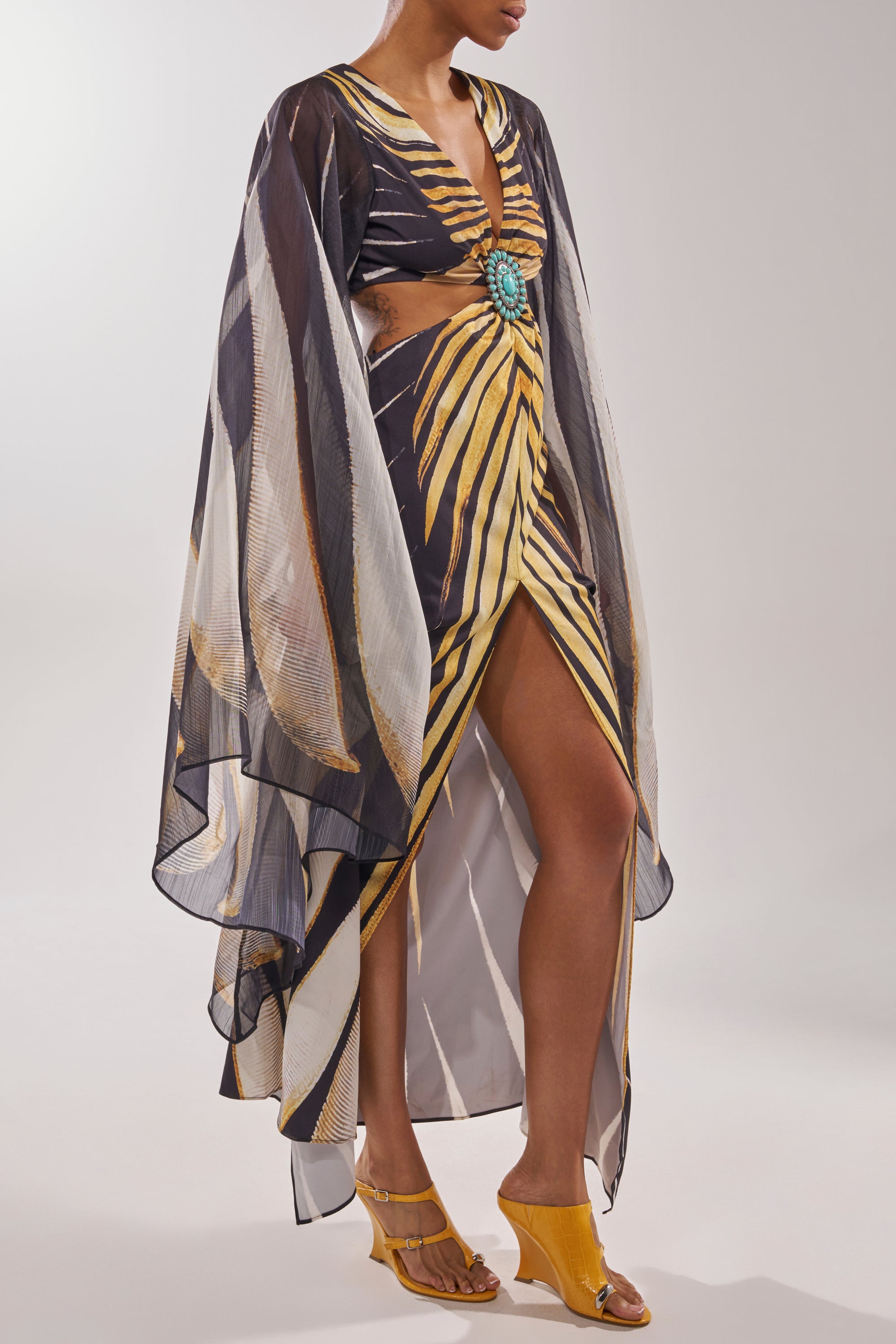 A woman, her face unseen, pairs a flowing, patterned dress with wide sheer sleeves and a deep V neckline with the AKIRA GOLD GINO MUSTARD CROC WEDGE SANDAL. A turquoise brooch at her waist completes the look. 1