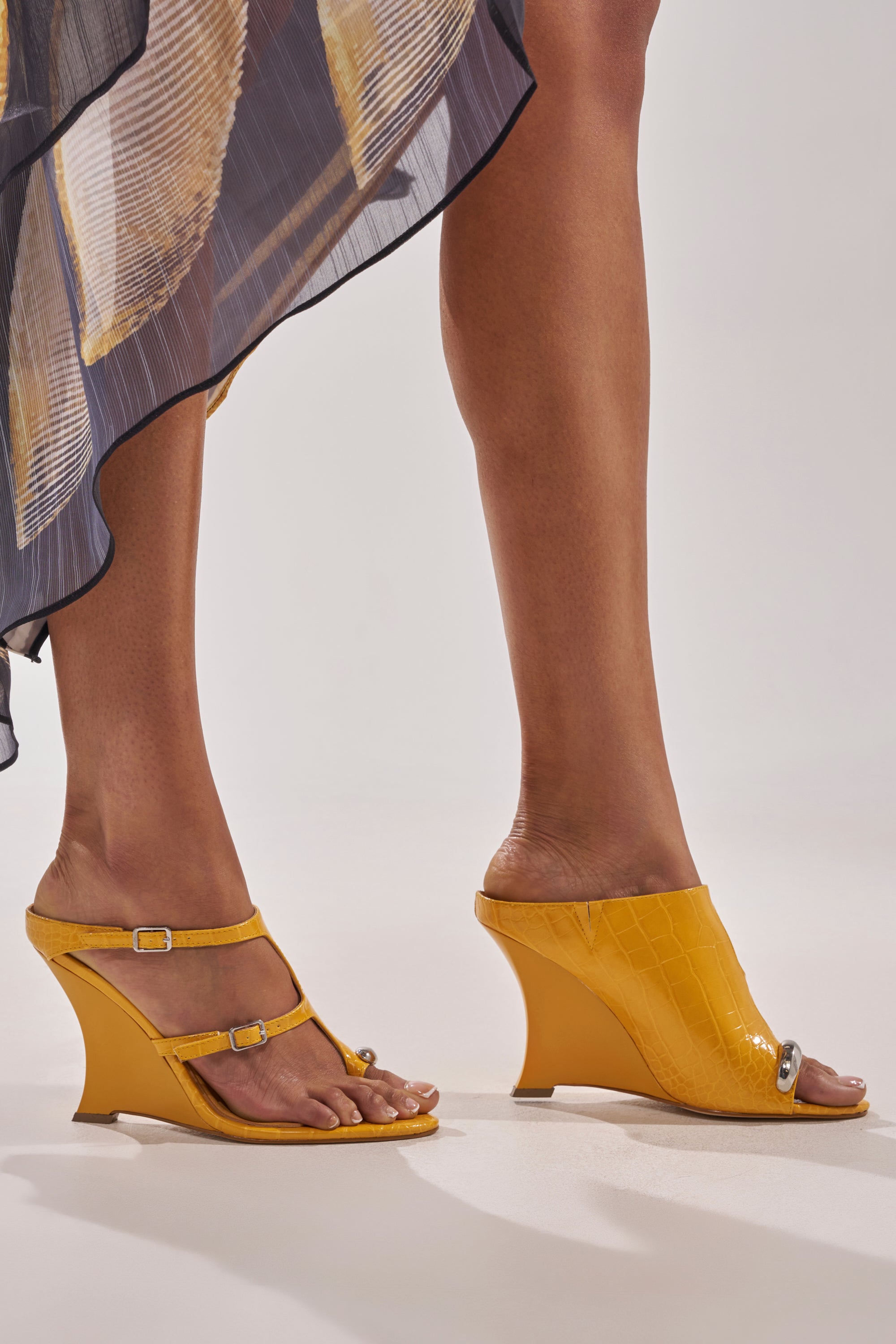 Wearing a sheer, patterned skirt, she pairs the AKIRA GOLD GINO MUSTARD CROC WEDGE SANDAL in a double-strap style on one foot and a closed mule on the other—both crafted from croc-embossed faux leather for comfy wedge appeal. 7
