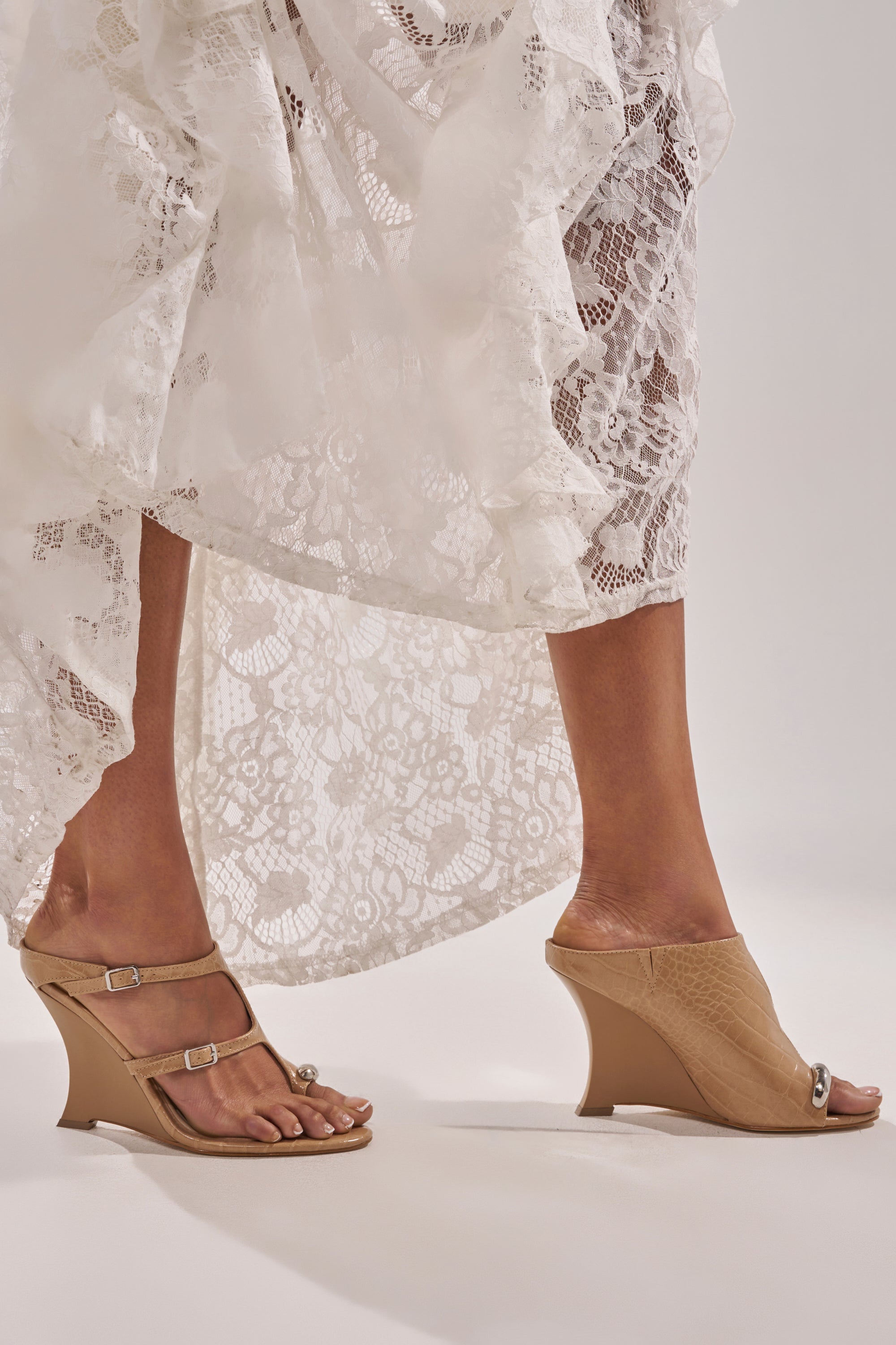 A person in a white lace dress stands with one foot in a tan double-strap sandal and the other in the AKIRA GOLD GINO NUDE CROC WEDGE SANDAL, both styles peeking out from beneath the hem. 0