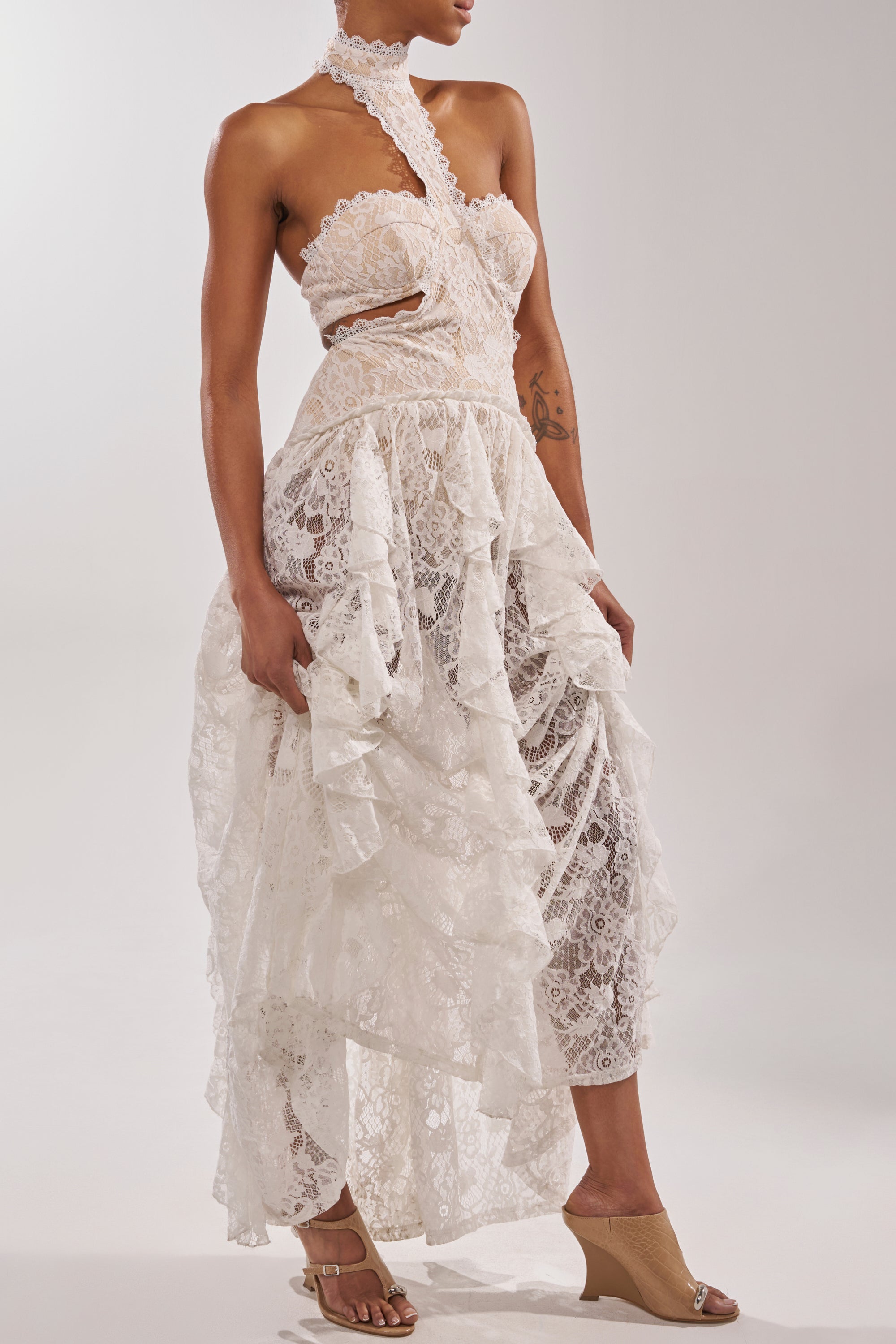 A woman in a white, asymmetrical lace dress with ruffles and a high neck strap stands against a plain light background, wearing the AKIRA GOLD GINO NUDE CROC WEDGE SANDAL for a stylish, comfortable look. 1