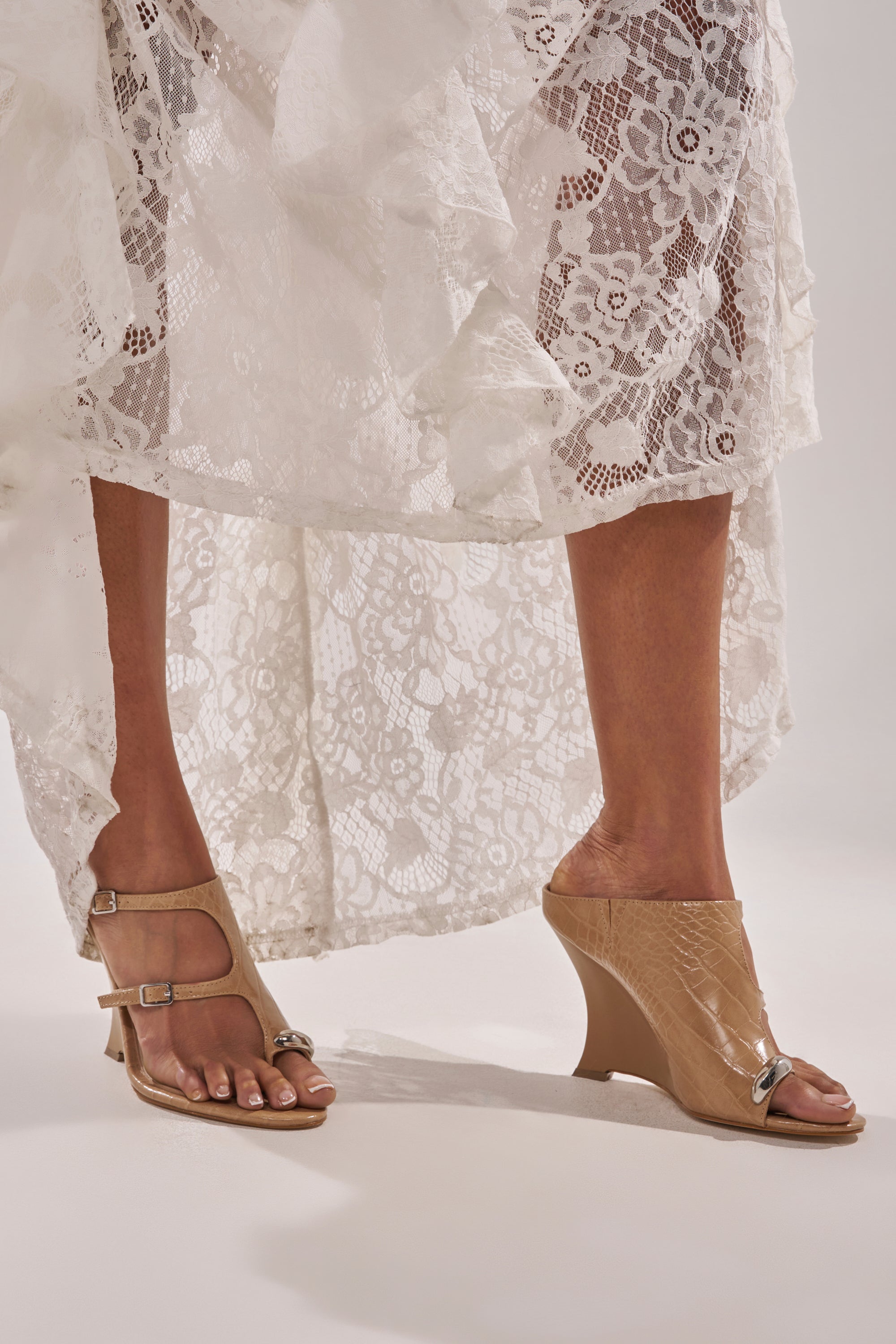 A person in a white lace skirt wears mismatched shoes: the right foot sports the AKIRA GOLD GINO NUDE CROC WEDGE SANDAL, while the left has a flat, faux croc-leather sandal with buckles. 2