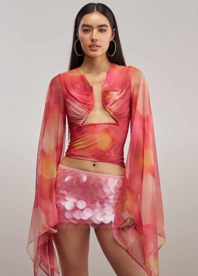 A model wearing a vibrant pink and orange tie-dye sheer long-sleeve crop top with a cutout, paired with a matching pink sequined mini skirt, against a solid light gray background.