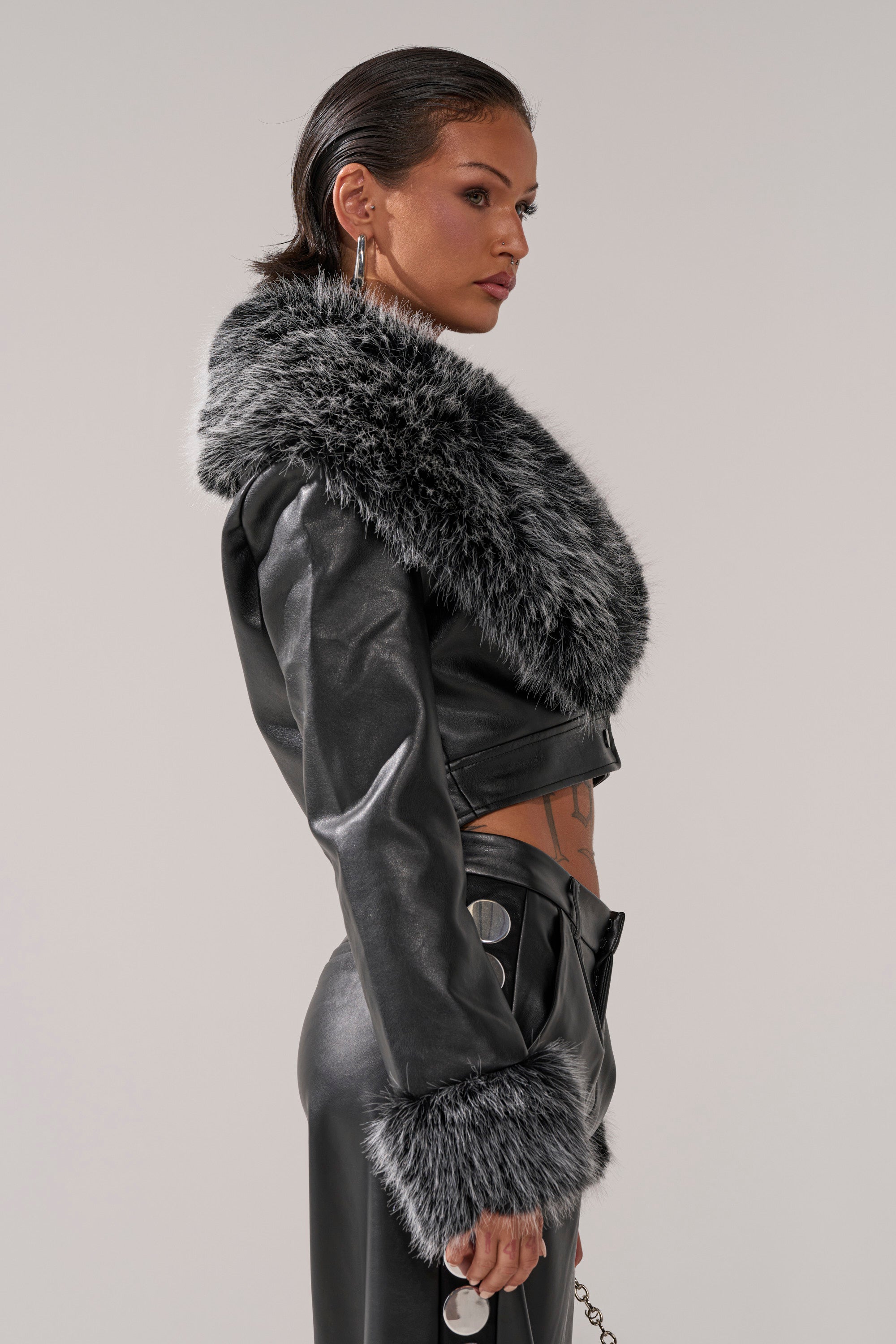 A woman in profile wears the GISELE CROP MOTO BLAZER IN BLACK GREY, styled with silver hoop earrings and button-detailed pants. Her hair is slicked back as she stands confidently against a plain background.