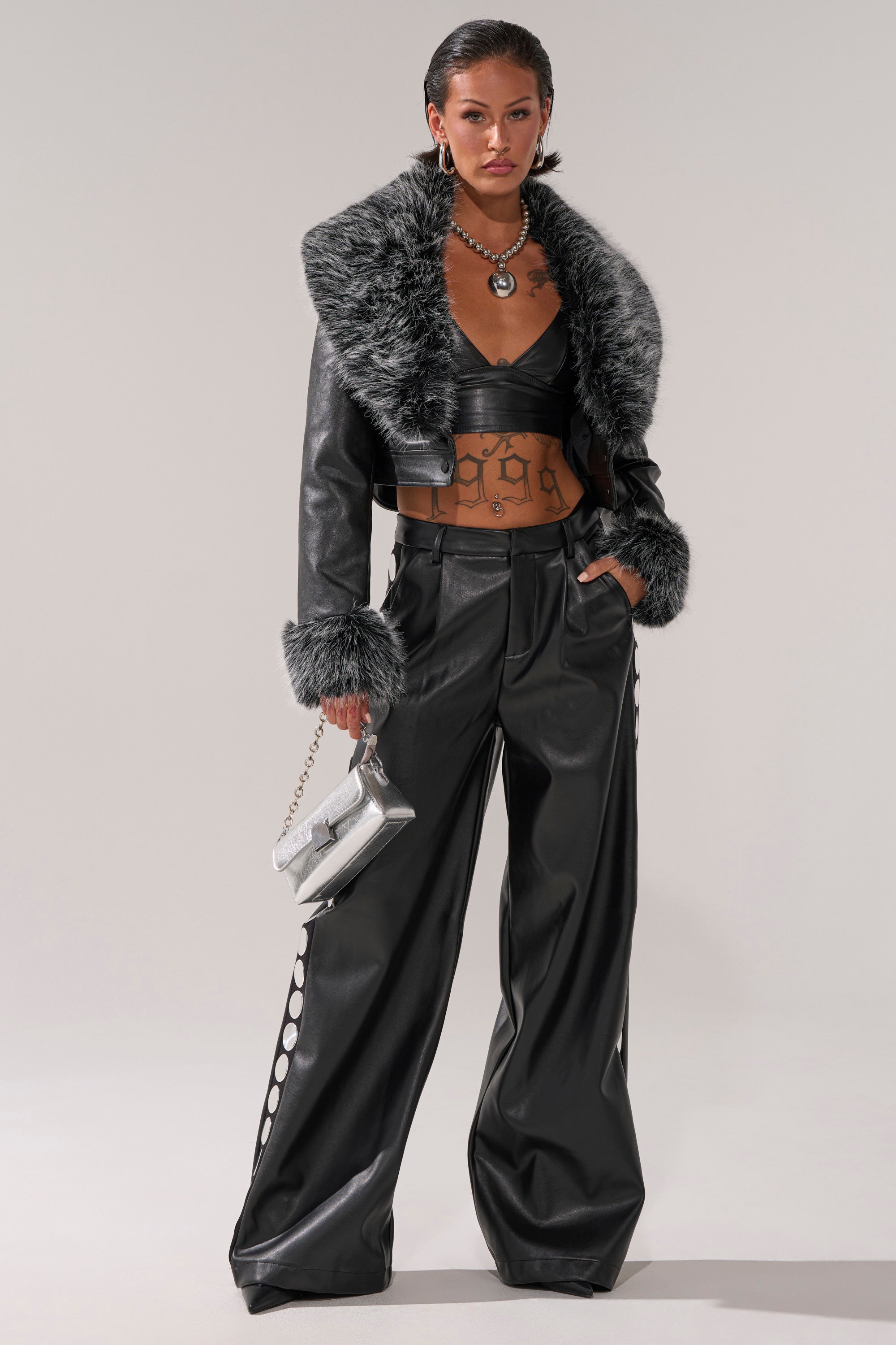 A woman exudes confidence in the GISELE CROP MOTO BLAZER IN BLACK GREY, paired with wide-leg pants, a crop top, bold jewelry, and a metallic handbag.