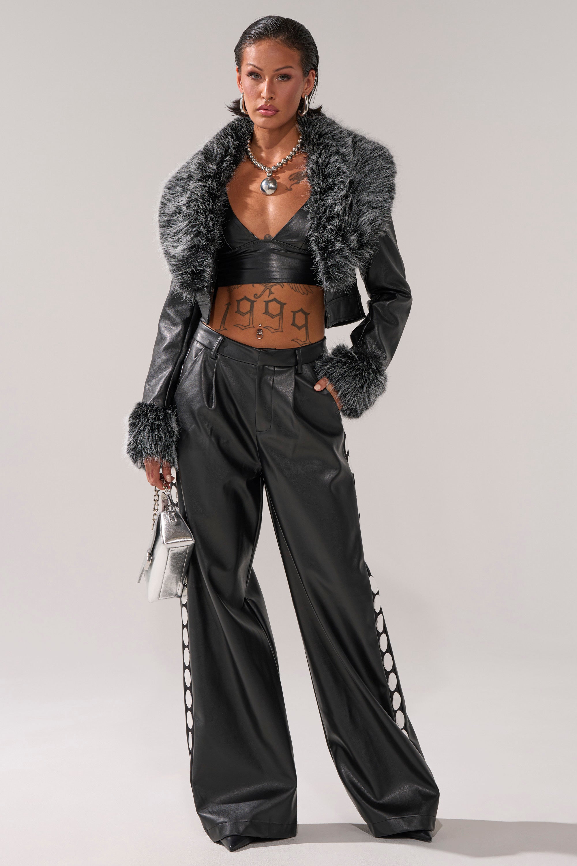 A woman wears the GISELE CROP MOTO BLAZER IN BLACK GREY with a black faux leather crop top, wide-leg pants with white cut-out details, silver jewelry, a metallic silver purse, and slicked-back hair.