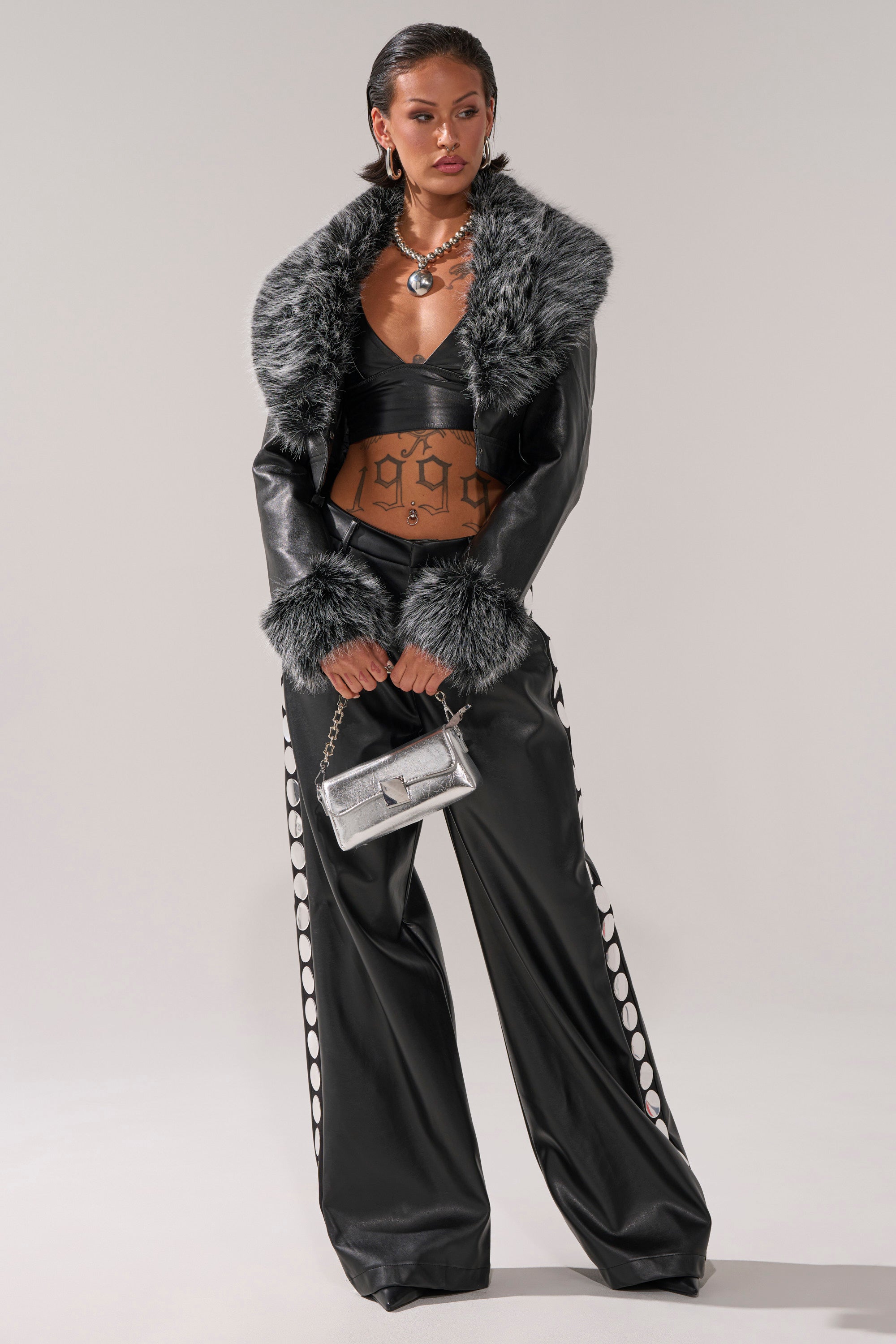 Wearing the GISELE CROP MOTO BLAZER IN BLACK GREY, wide-leg pants with circular cutouts, a silver necklace, earrings, and holding a small metallic purse, a person poses confidently against a plain background.