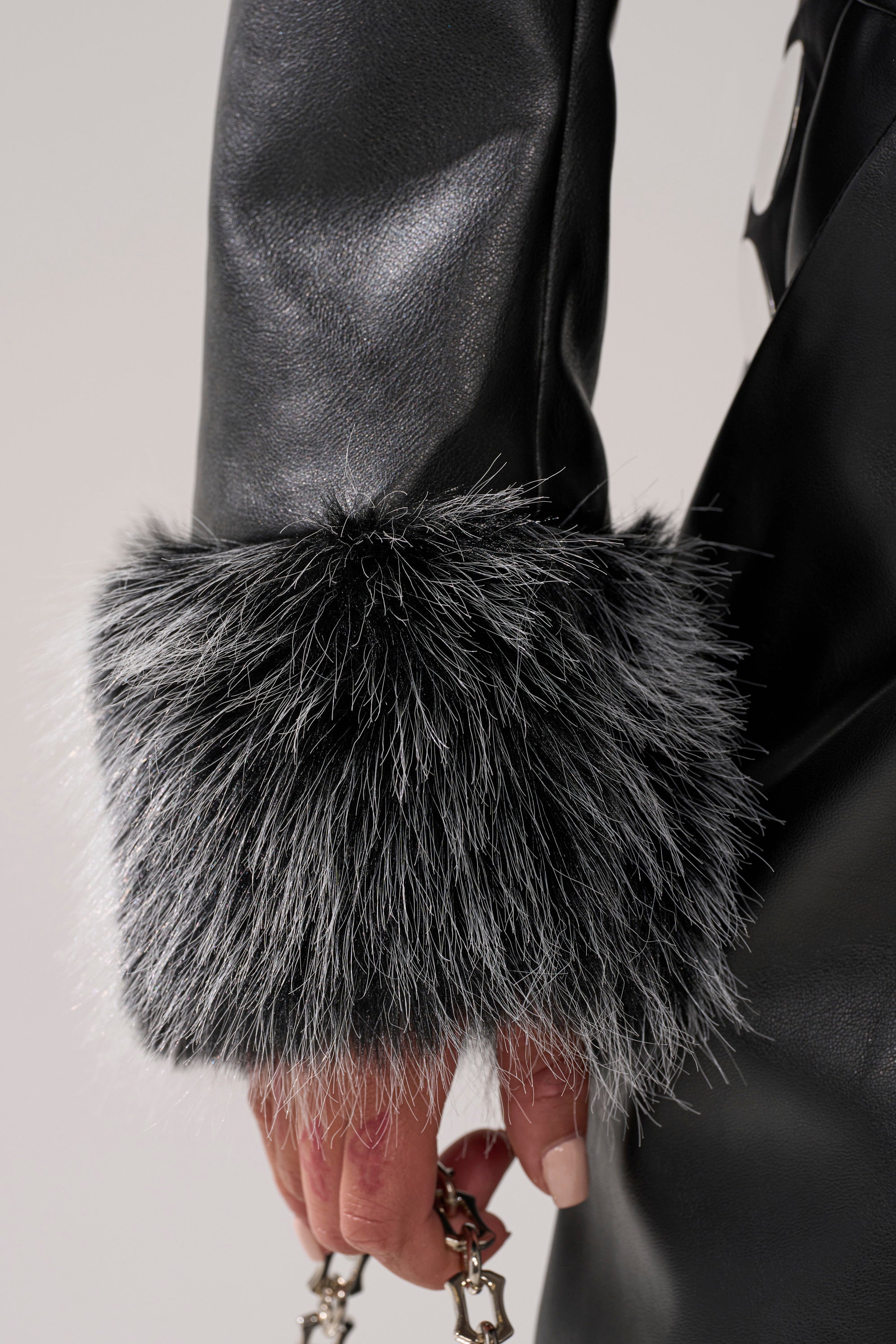 A close-up shows someone wearing the GISELE CROP MOTO BLAZER IN BLACK GREY with fuzzy black-and-white faux fur cuffs, holding a silver chain accessory.