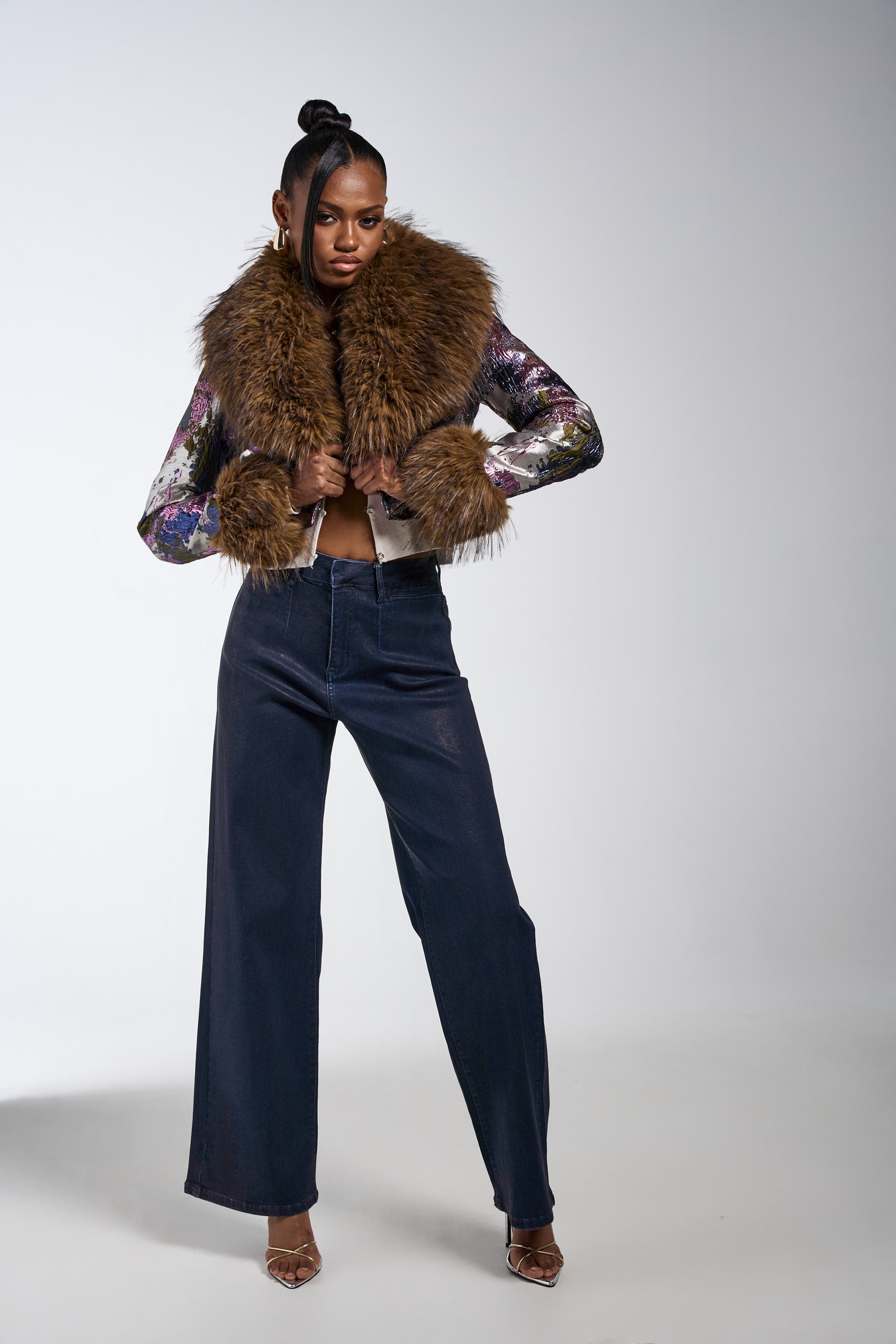 A woman stands confidently in the GISELE FUR TRIM MOTO IN BROCADE jacket—featuring colorful sleeves and a faux fur collar—paired with wide-leg dark jeans and clear heels, set against a plain white background. 9