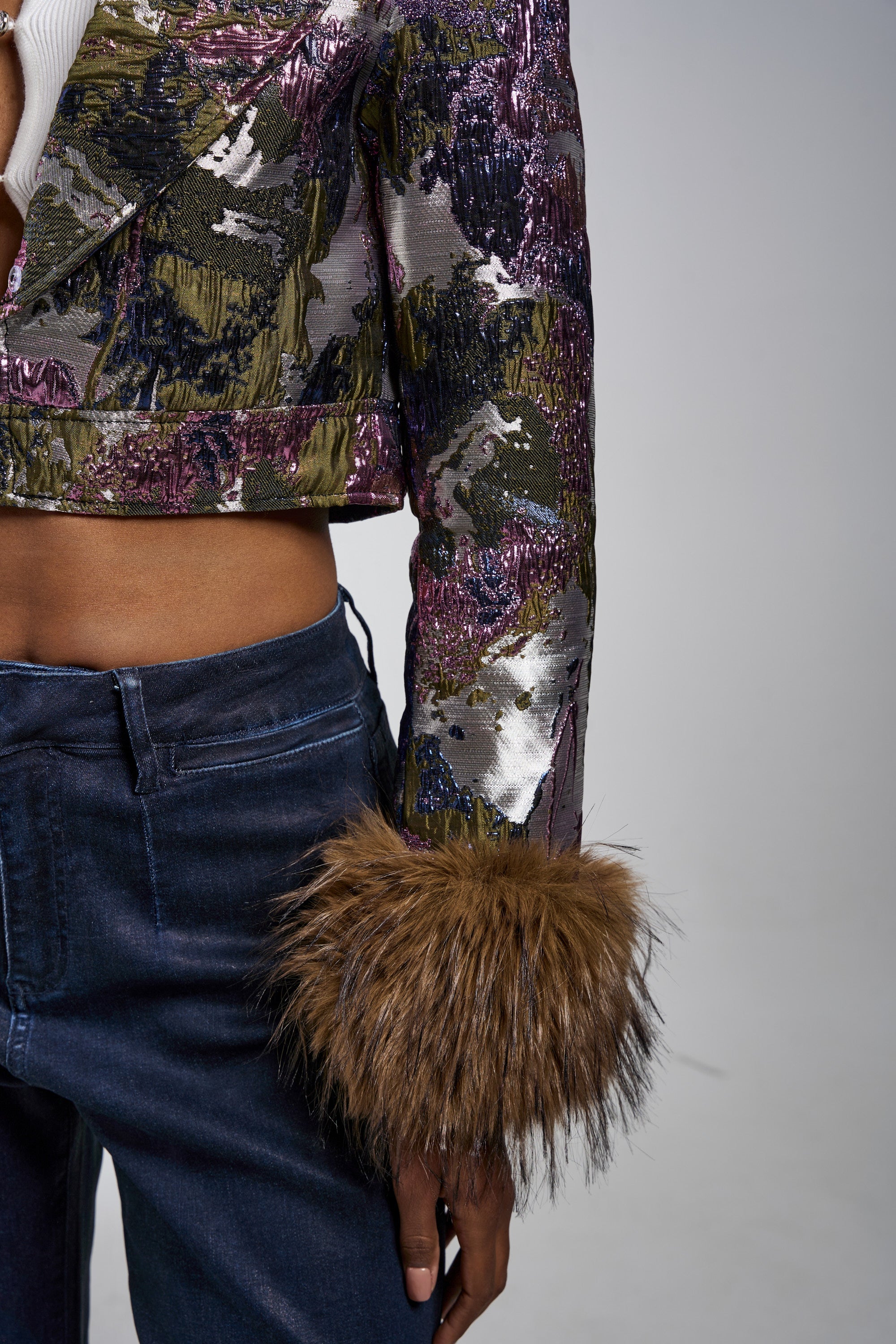 Wearing the GISELE FUR TRIM MOTO IN BROCADE, a person's torso and left arm are shown; the jacket features multicolored metallic brocade with a furry brown cuff, paired with dark blue jeans against a plain background. 10