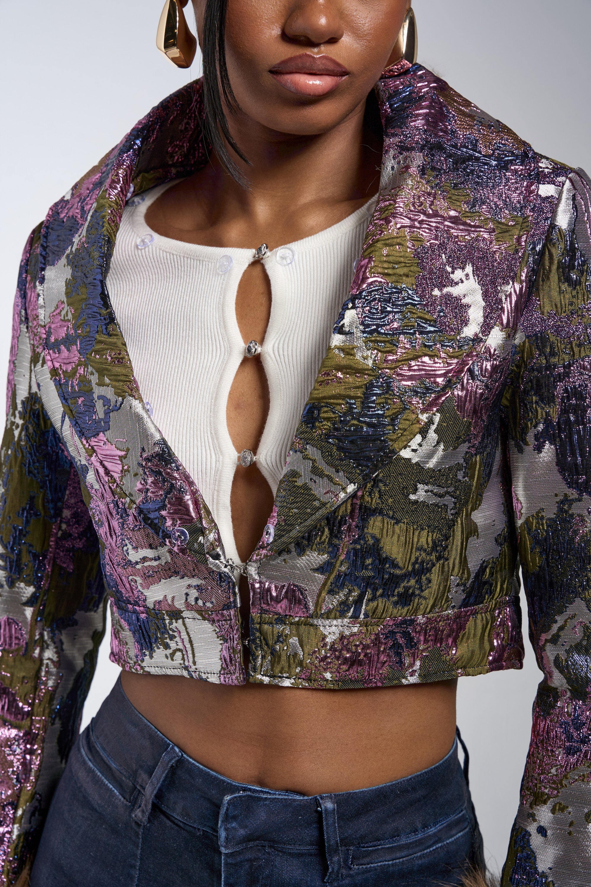 A woman wears the GISELE FUR TRIM MOTO IN BROCADE jacket over a white ribbed top with jeweled buttons and dark high-waisted jeans, showing only her lower face, torso, and one ear with an earring. 11