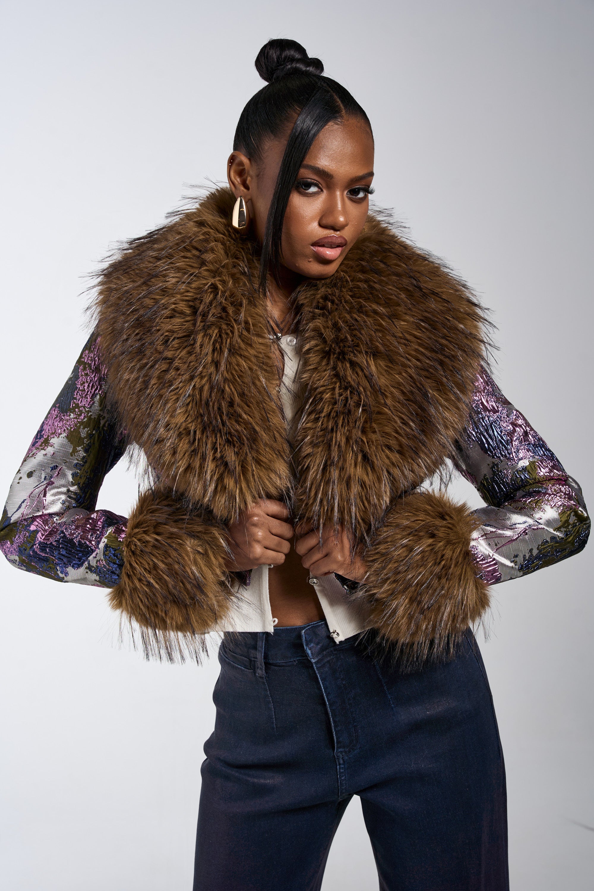 A woman with a sleek top bun poses confidently in the GISELE FUR TRIM MOTO IN BROCADE, a metallic brocade jacket with faux fur collar and cuffs, styled with dark pants against a plain background. 1