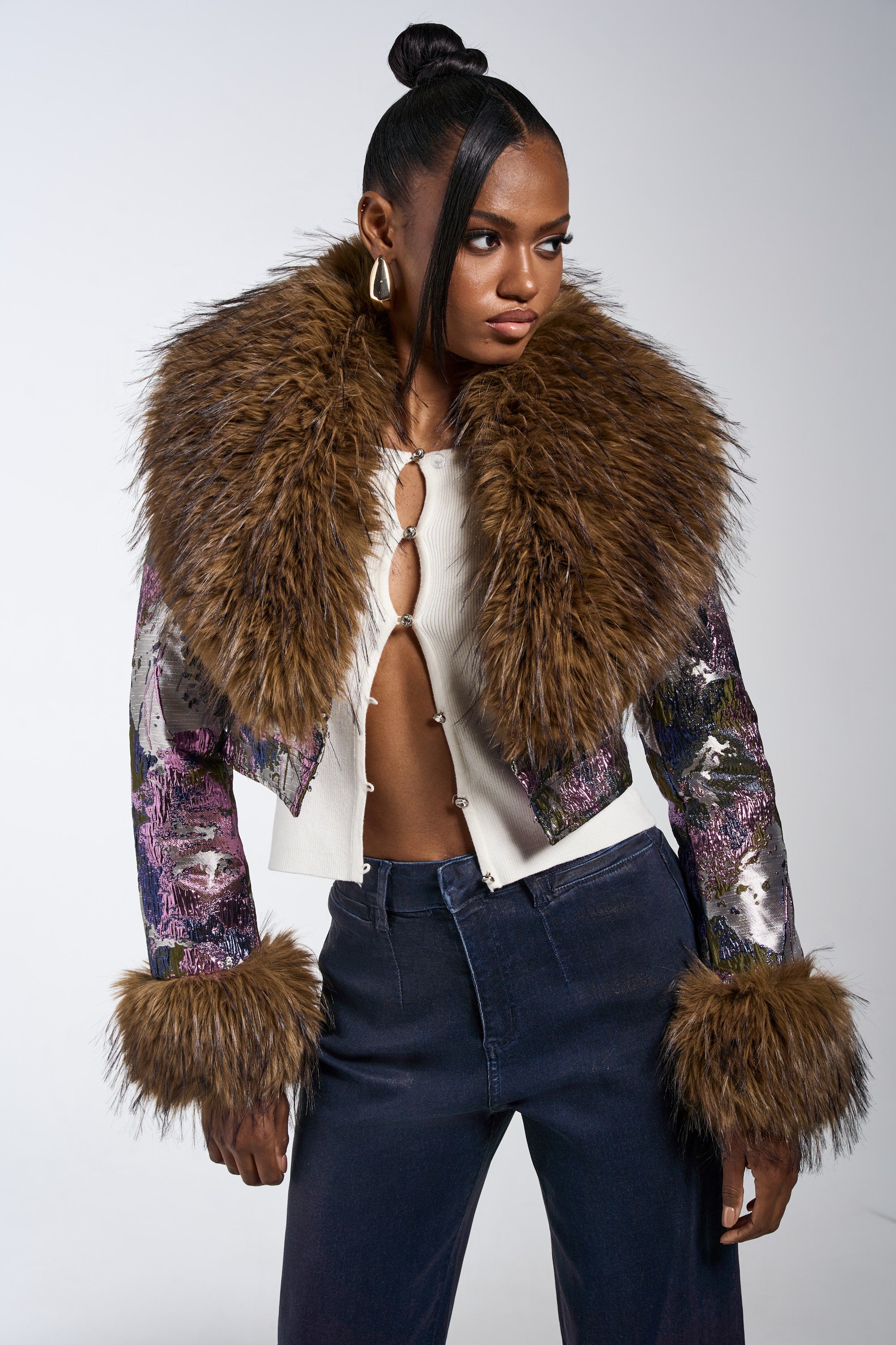 A woman with a high bun wears the GISELE FUR TRIM MOTO IN BROCADE—a metallic brocade jacket with a large brown faux fur collar and cuffs—over a cream top and navy pants, standing hands on hips against a plain background. 2