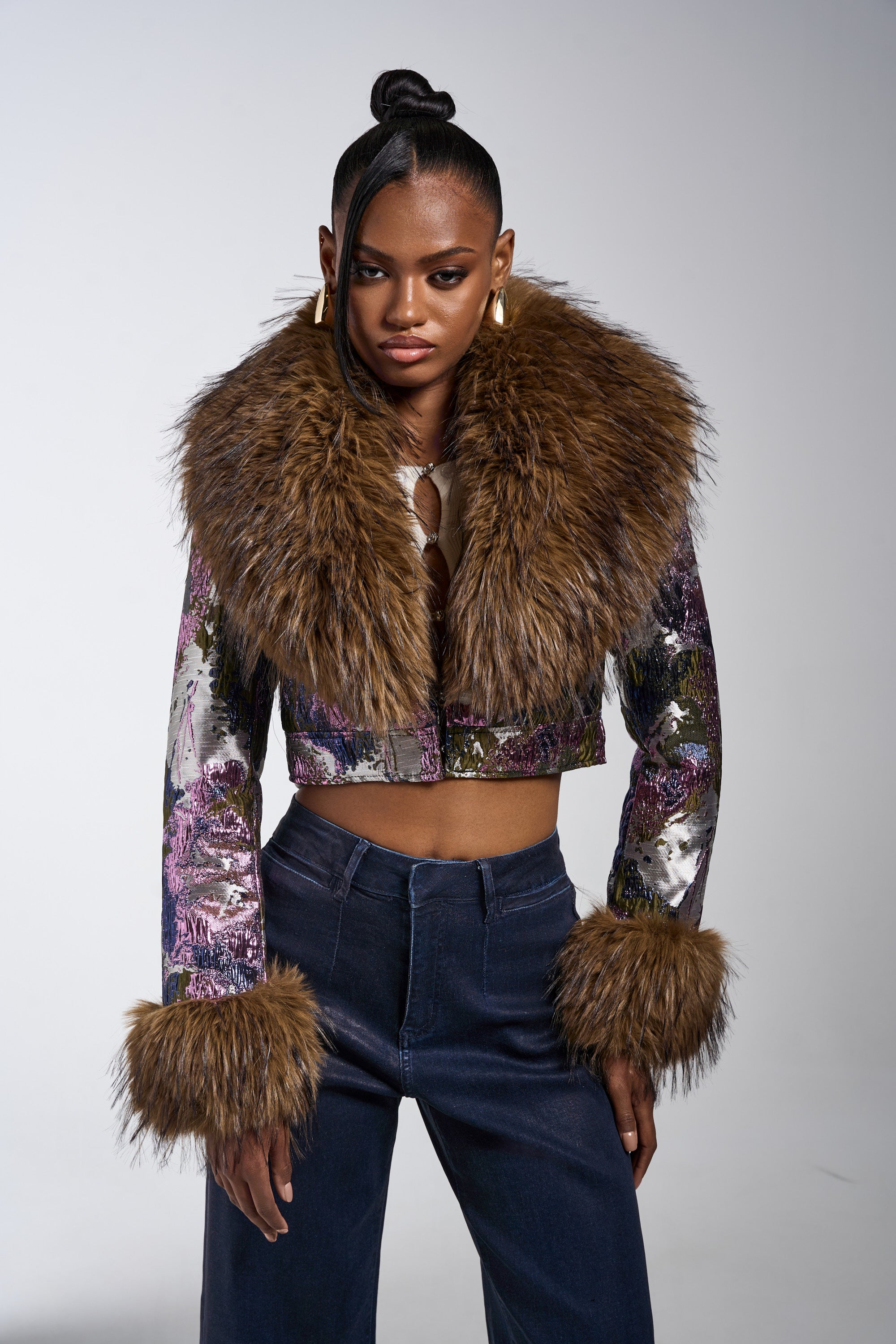 A woman exudes confidence in the GISELE FUR TRIM MOTO IN BROCADE, featuring a bold faux fur collar and cuffs, styled with high-waisted dark blue pants, gold earrings, and a sleek hairstyle. 4
