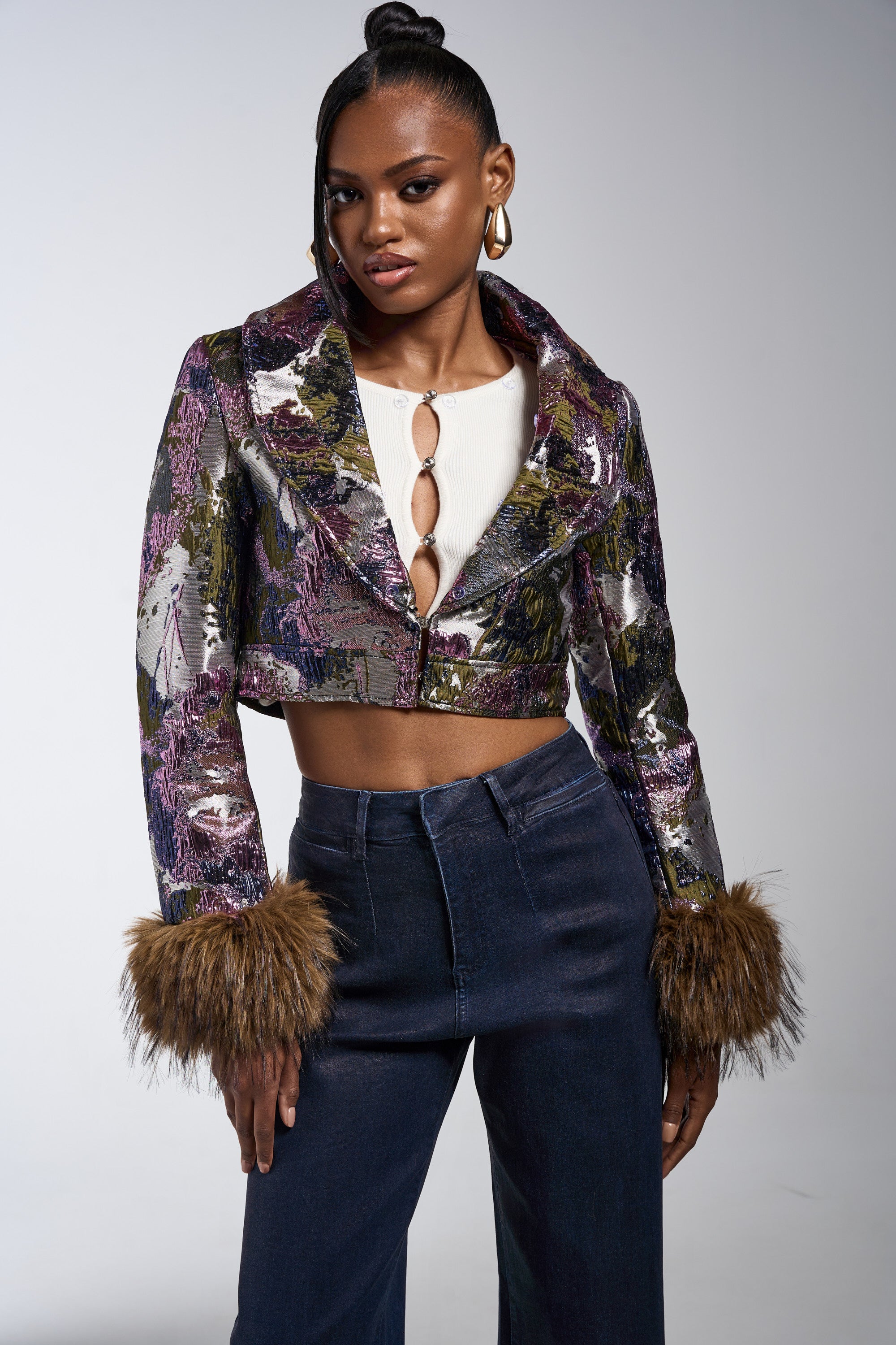 A woman poses confidently in the GISELE FUR TRIM MOTO IN BROCADE jacket with furry cuffs, styled over a white top with decorative rings and high-waisted dark jeans. She accessorizes with hoop earrings and wears her hair in a high bun. 6