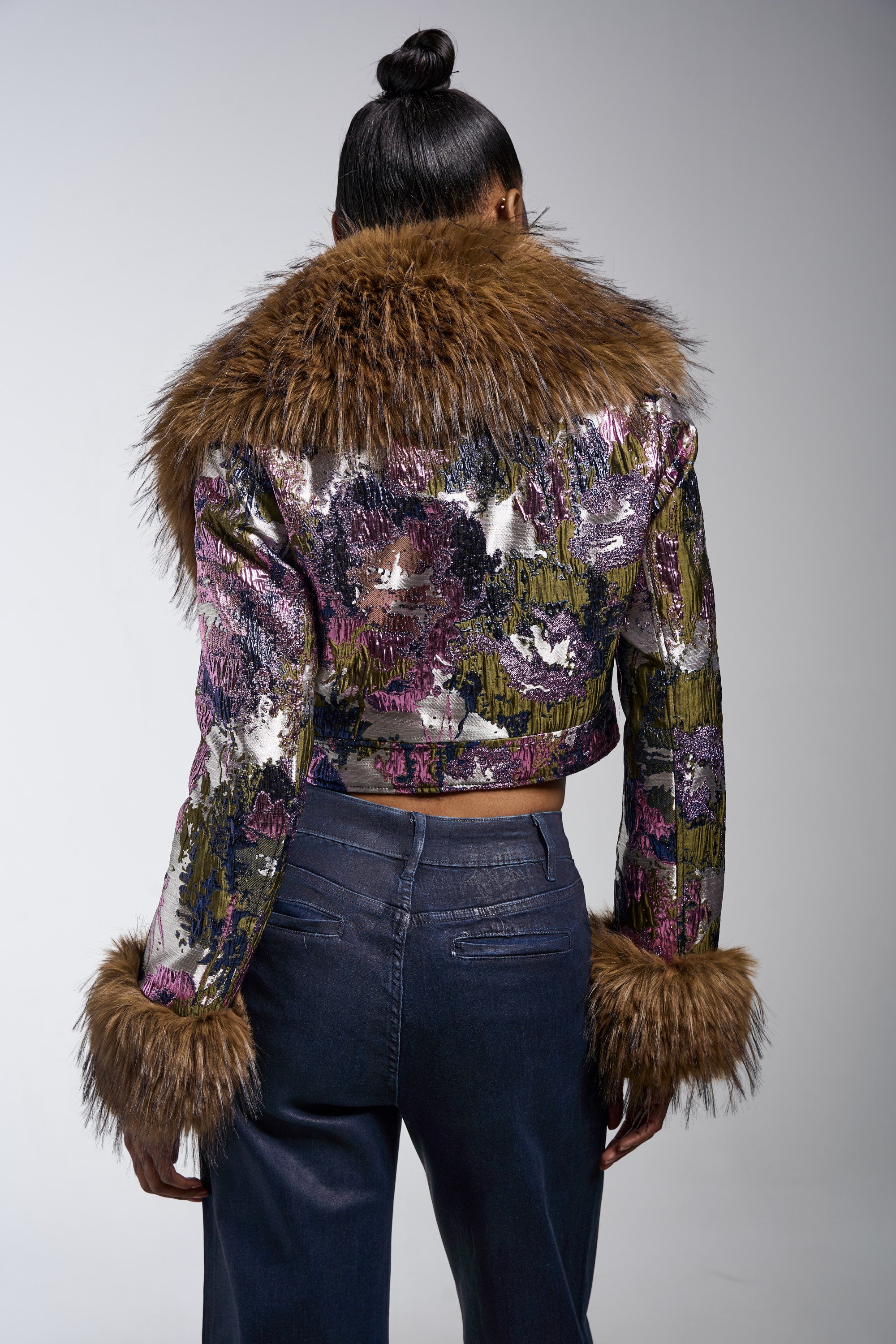 A person with their hair in a bun is seen from behind, wearing the GISELE FUR TRIM MOTO IN BROCADE jacket with a faux fur collar and cuffs, paired with high-waisted dark jeans. 8