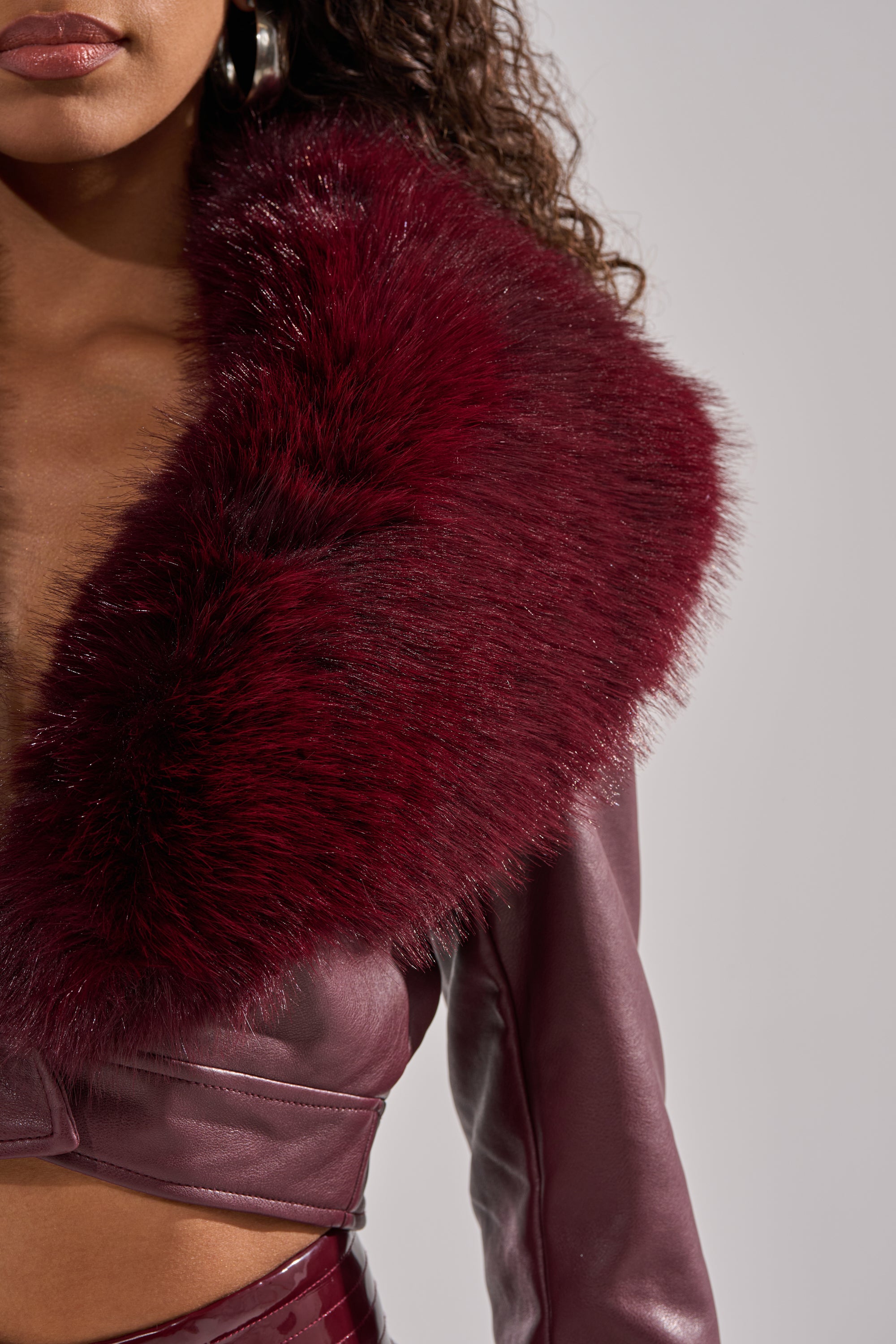 A woman models the GISELE FUR TRIM MOTO IN BURGUNDY, featuring deep burgundy faux leather and a matching oversized faux fur collar, shown from the shoulders down against a neutral background.