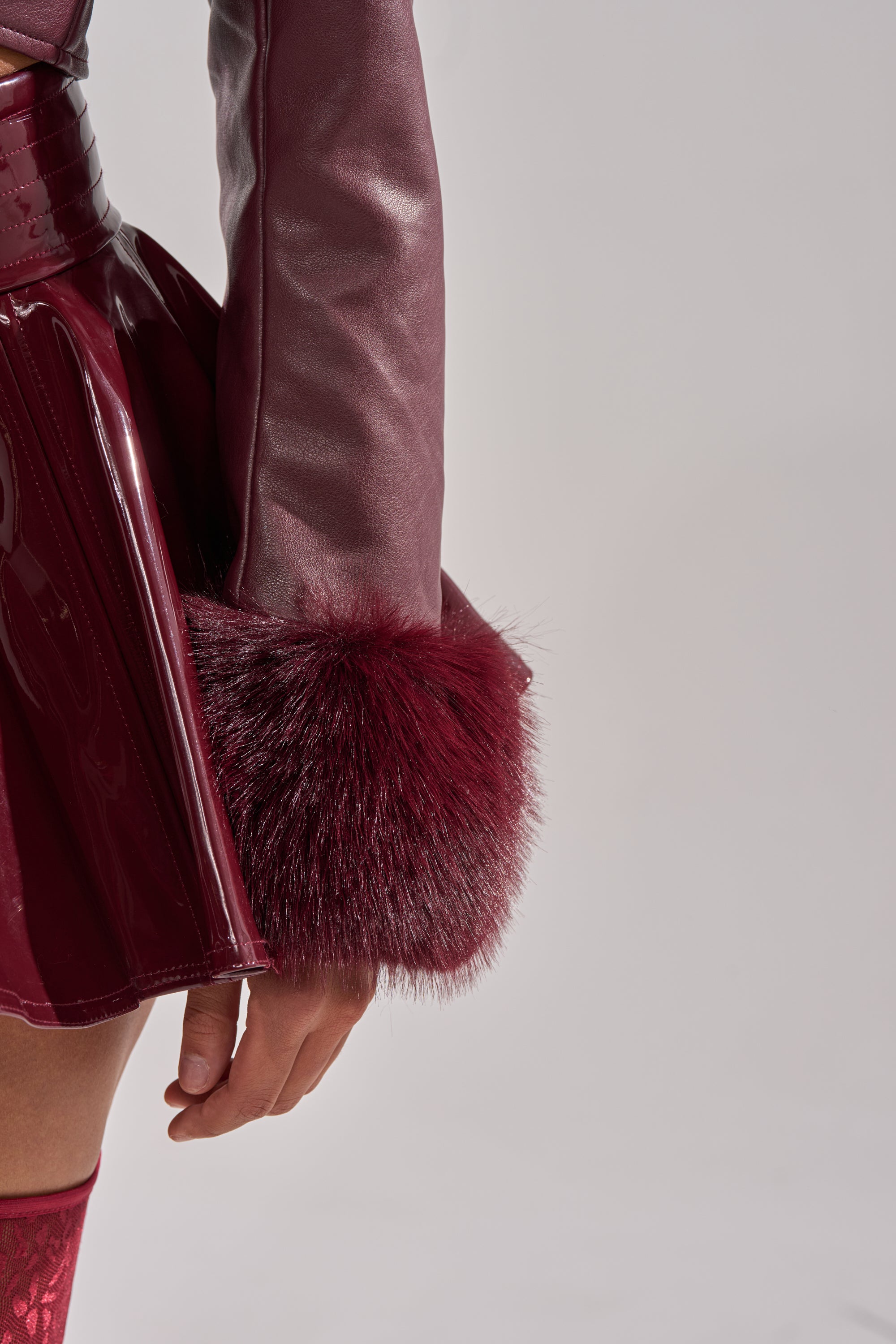 A person models the GISELE FUR TRIM MOTO IN BURGUNDY, a faux leather jacket with fur trim cuffs, paired with a shiny matching skirt, their arm relaxed at their side against a light gray background.