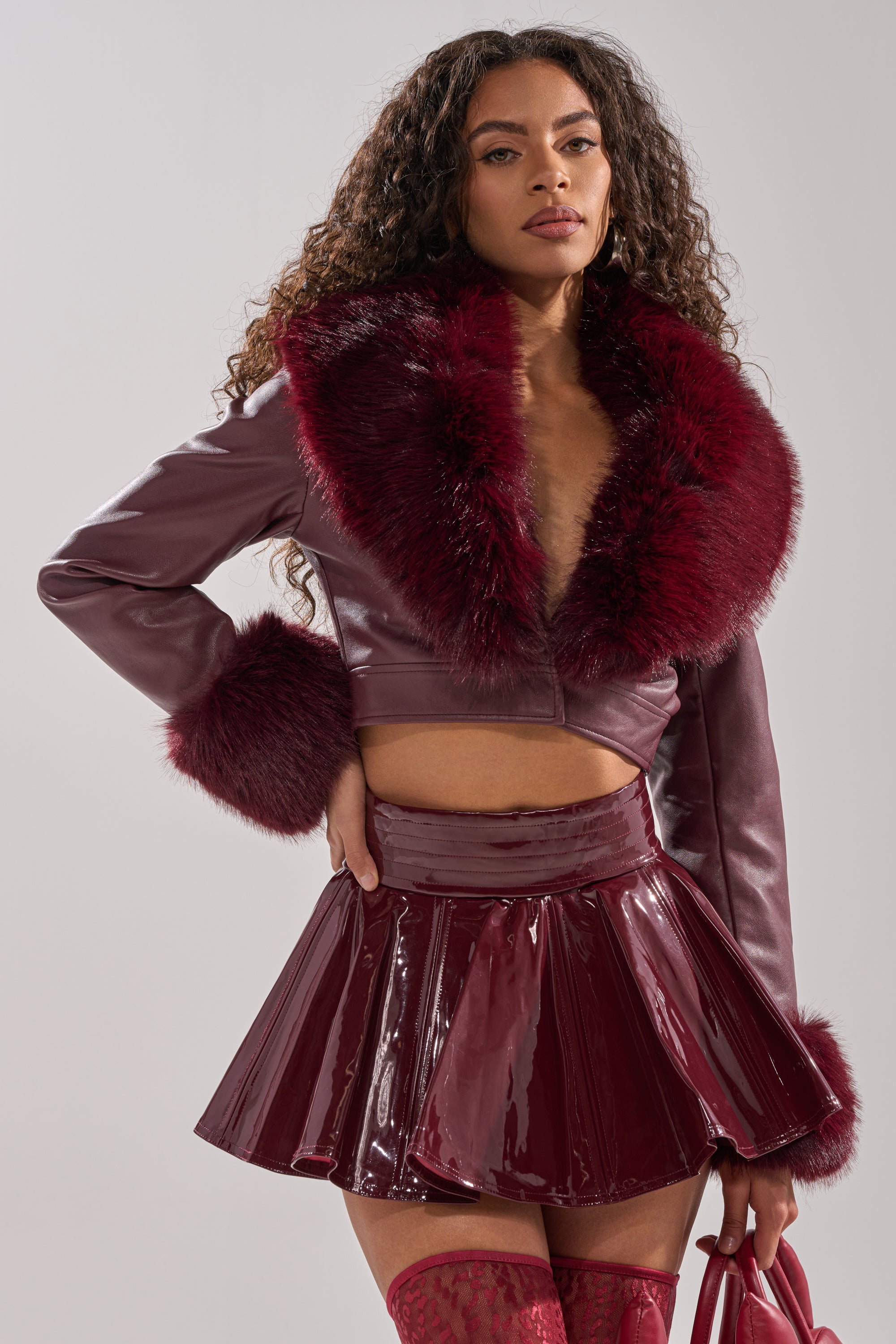 A woman with long curly hair wears the GISELE FUR TRIM MOTO IN BURGUNDY and a shiny matching mini skirt. She holds a small handbag and gazes confidently at the camera against a neutral background.