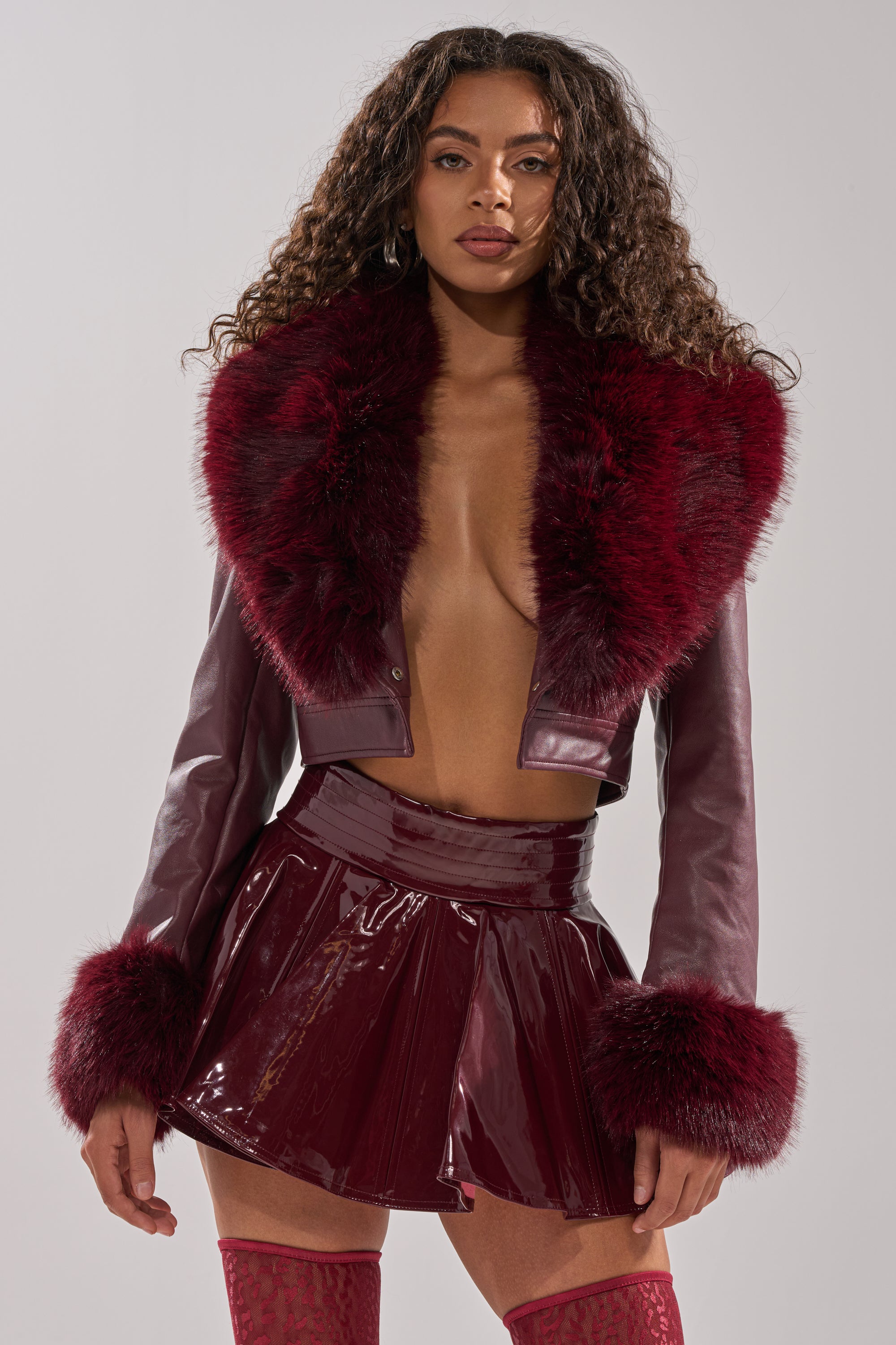 A woman with curly hair models the GISELE FUR TRIM MOTO IN BURGUNDY with faux fur collar and cuffs, paired with a shiny burgundy mini skirt and matching thigh-high stockings against a plain light background.