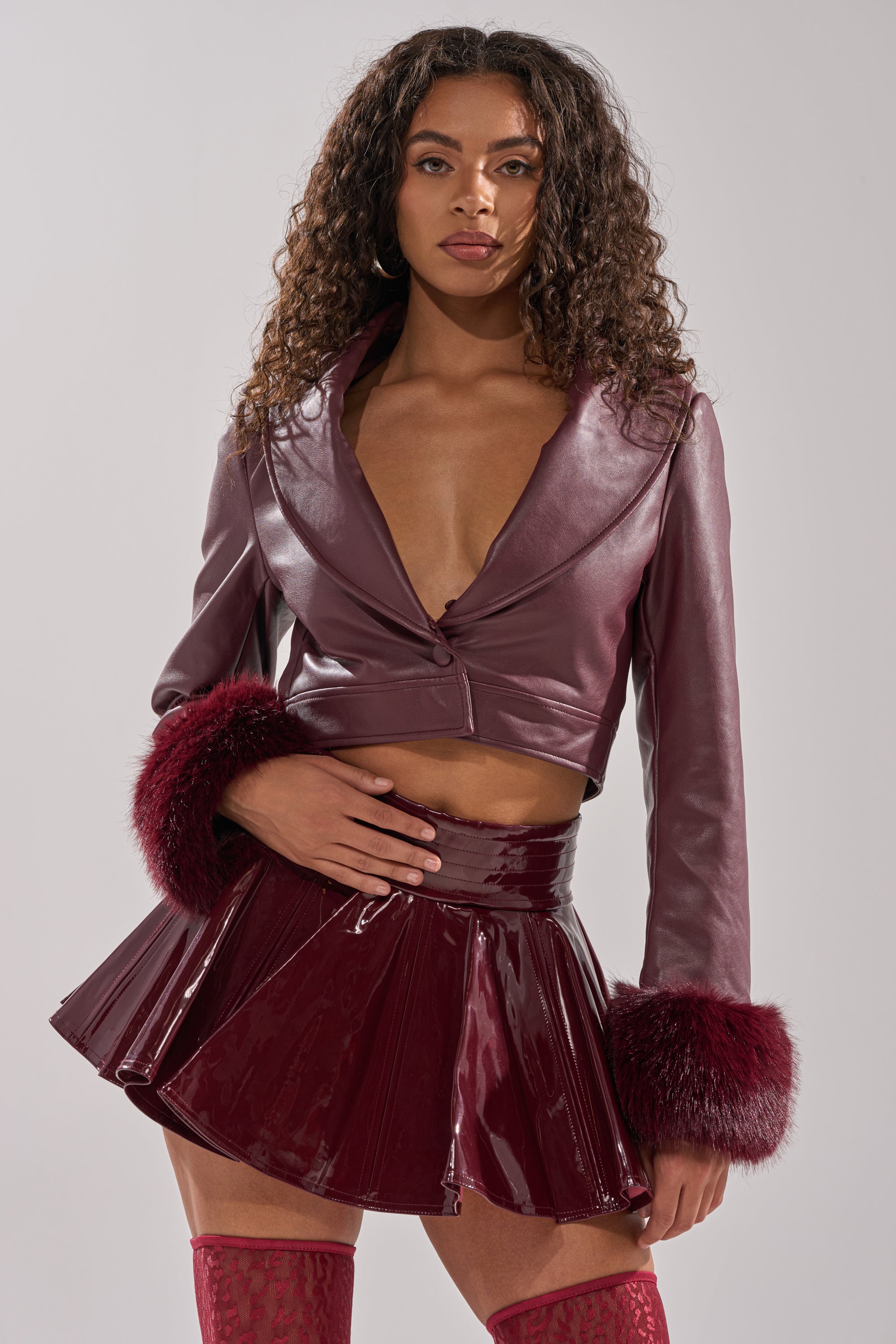 A woman with curly hair and a serious expression stands confidently in the GISELE FUR TRIM MOTO IN BURGUNDY, featuring faux fur cuffs, paired with a shiny pleated mini skirt against a plain background.