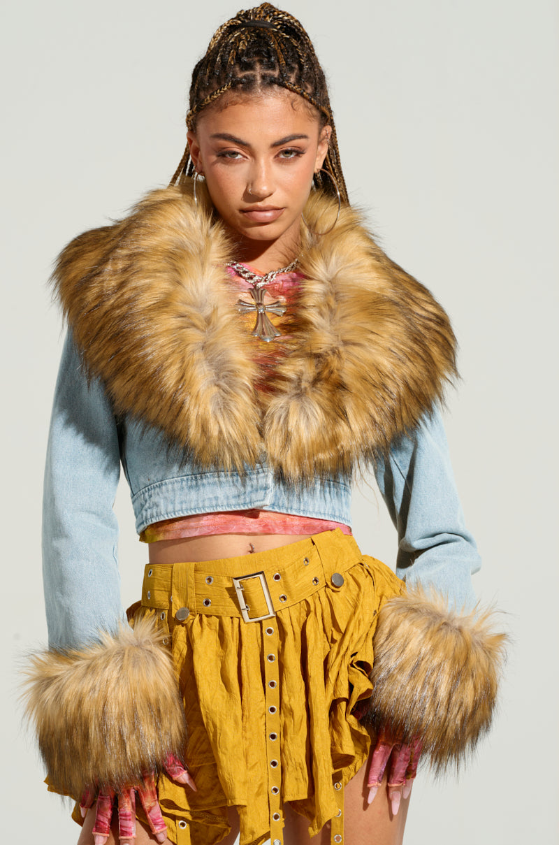 A model with braids poses in a stylish outfit featuring the NEW GISELE CROPPED FUR TRIM JACKET, paired with a yellow skirt adorned with ruffles and a belt. The light wash denim jacket flaunts prominent faux fur details on the collar and cuffs. The model stands against a plain, light-colored background, exuding Y2K revival vibes.