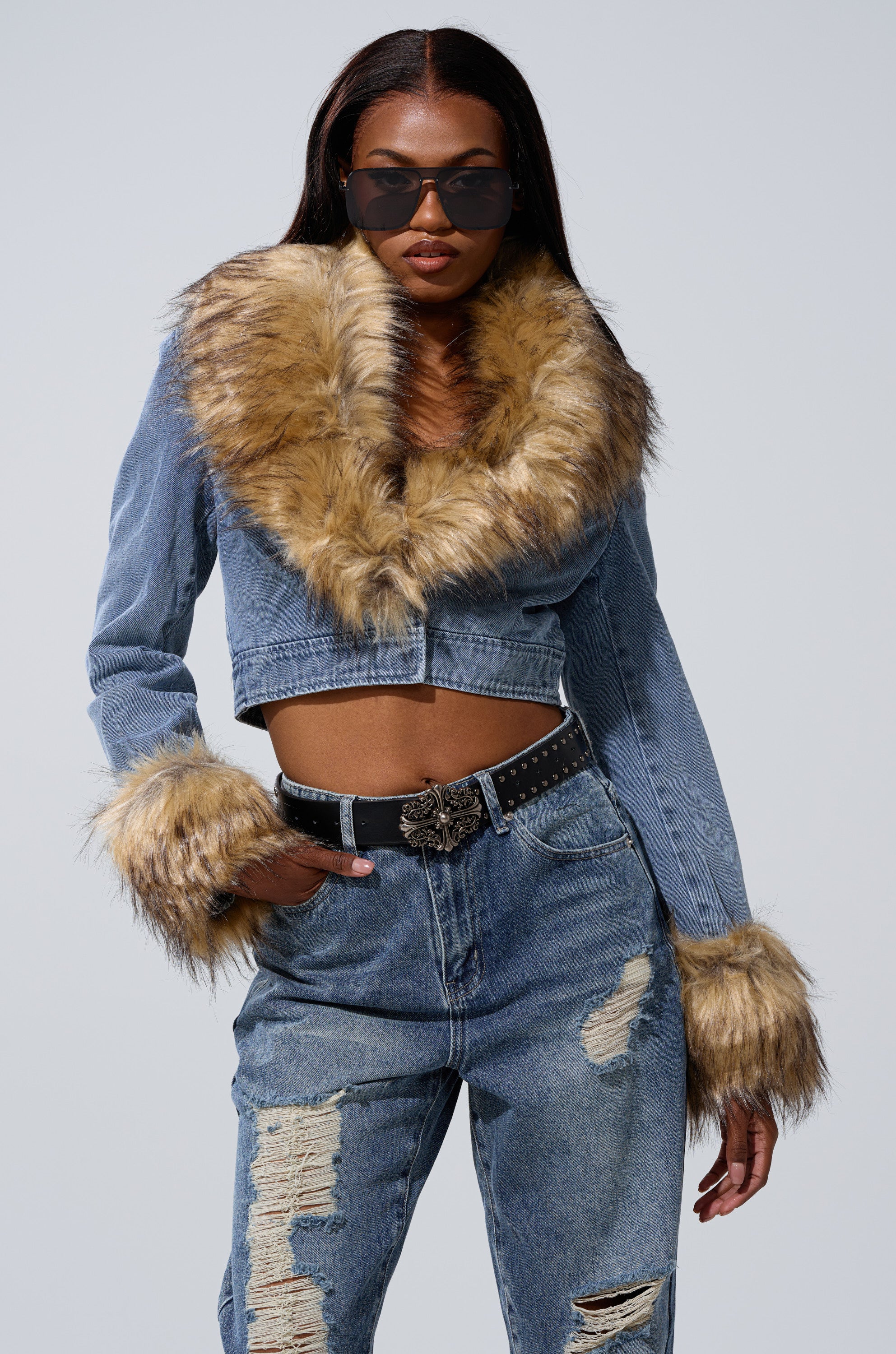 A woman in sunglasses, wearing the NEW GISELE CROPPED FUR TRIM JACKET with large faux fur details, a studded belt, and ripped high-waisted jeans stands confidently against a plain background.