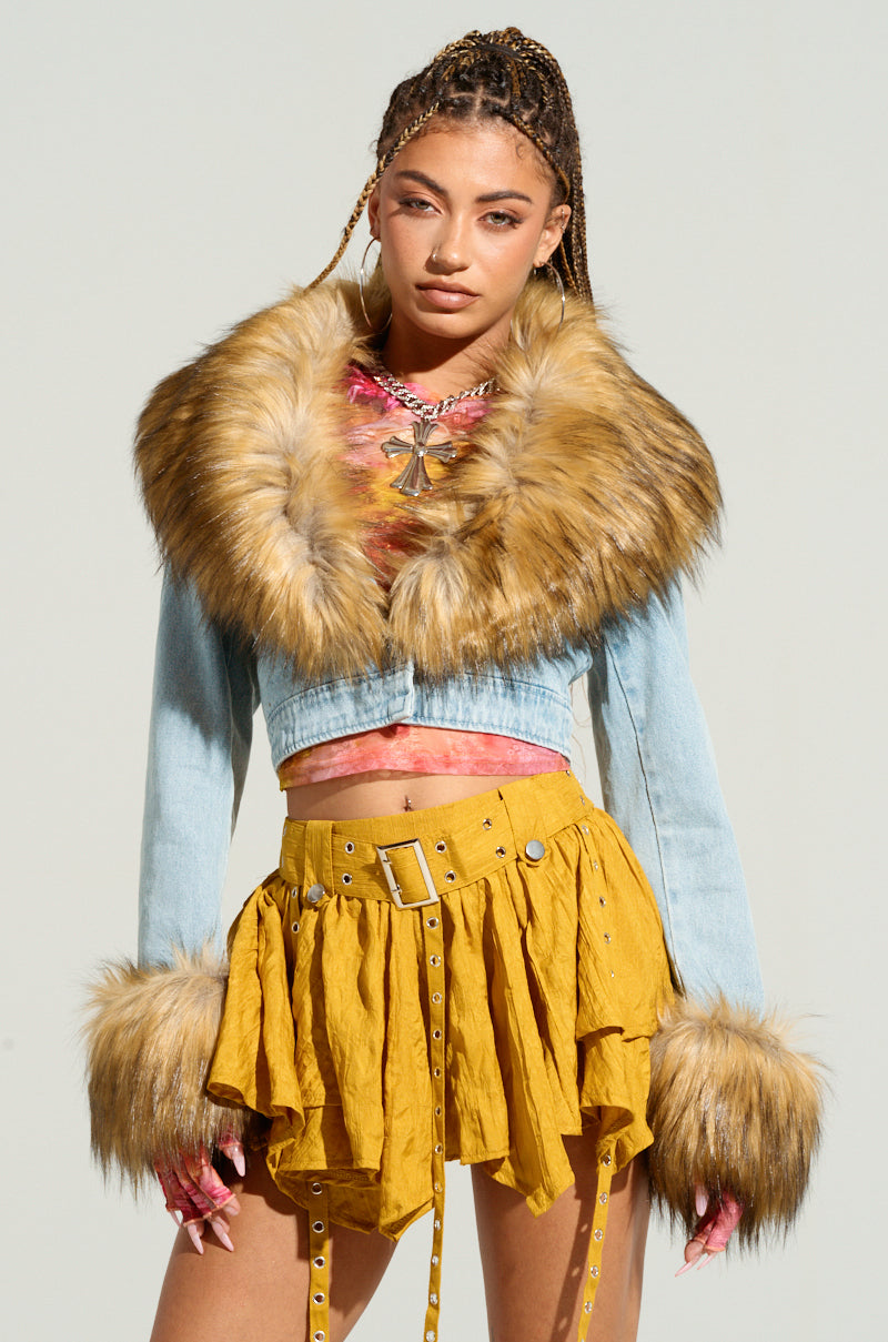 A person stands confidently, wearing the NEW GISELE Cropped Fur Trim Jacket in a light wash denim, perfectly paired with a mustard yellow, belted, layered mini skirt. They have long braided hair adorned with beads and sport a colorful top underneath the jacket, epitomizing Y2K revival fashion.