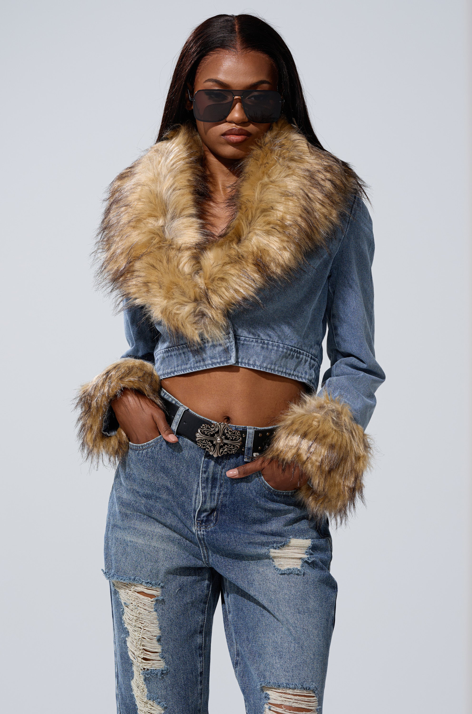 A woman wears sunglasses and the NEW GISELE CROPPED FUR TRIM JACKET featuring a large faux fur collar and cuffs, paired with ripped jeans. She poses confidently with one hand in her pocket against a plain background.