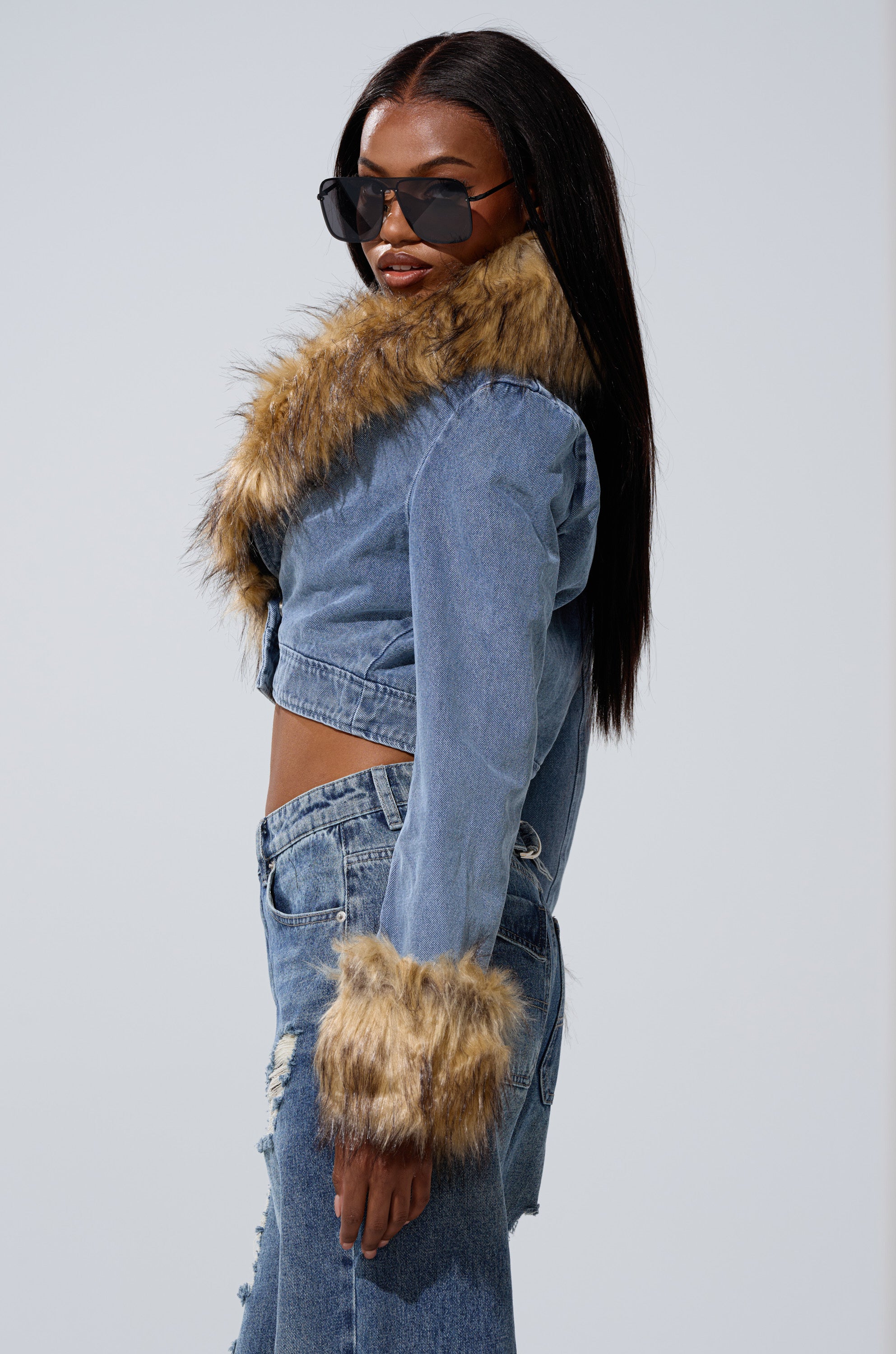 A woman in sunglasses models the NEW GISELE CROPPED FUR TRIM JACKET, featuring faux fur accents on the collar and cuffs, paired with ripped high-waisted jeans, posing confidently against a plain backdrop.