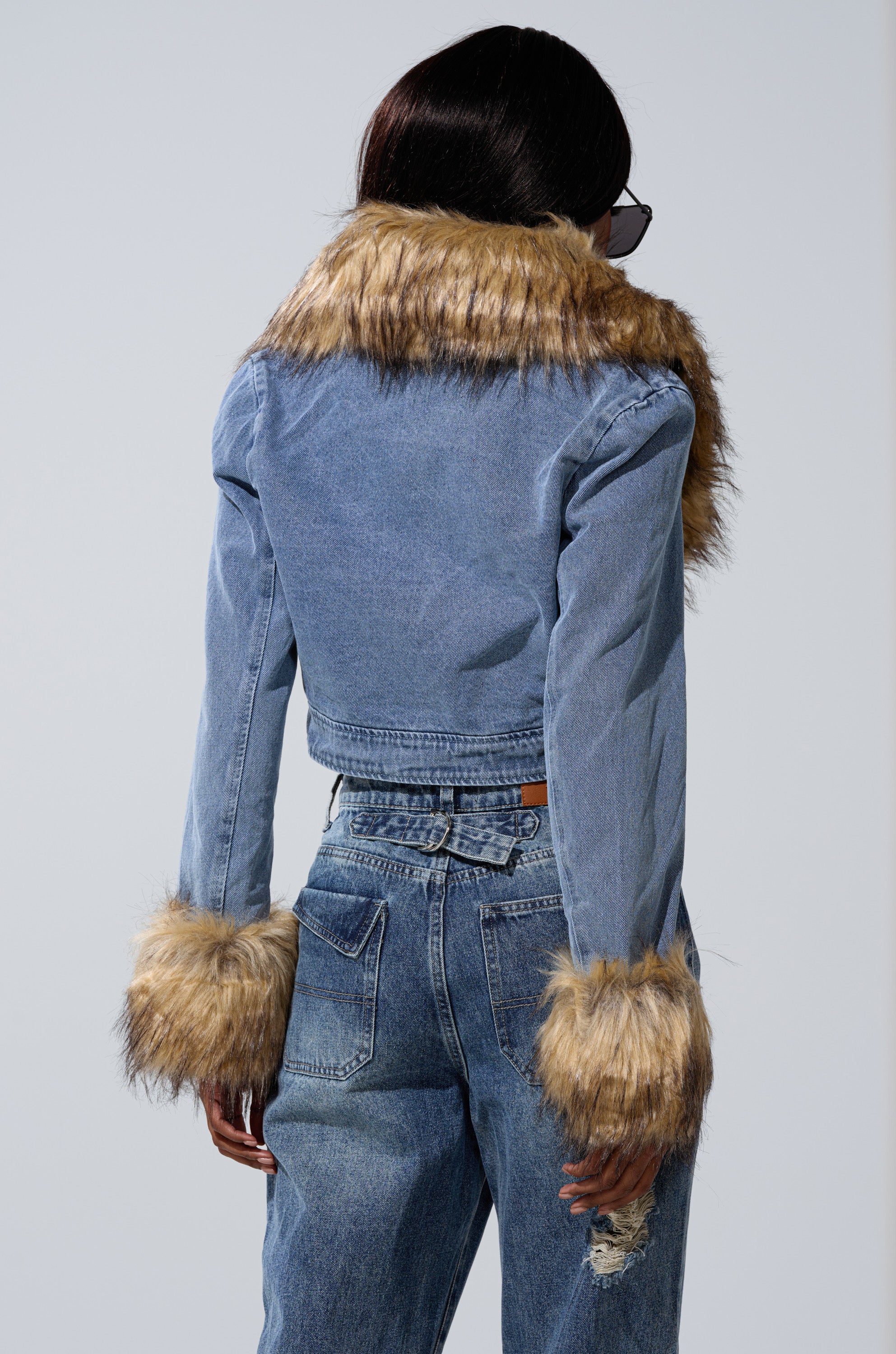 An individual faces away from the camera, wearing the NEW GISELE CROPPED FUR TRIM JACKET—a cropped denim jacket with faux fur collar and cuffs—styled with high-waisted blue jeans for a Bratz Doll-inspired look.