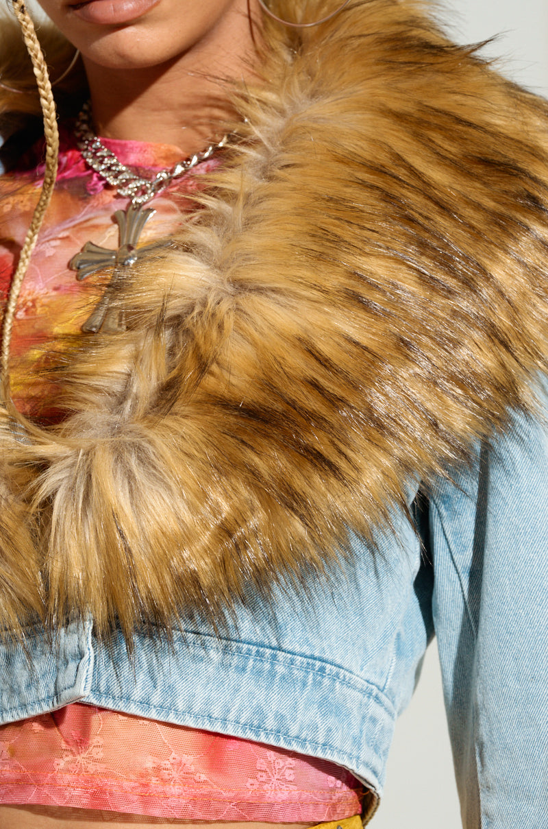 A close-up of a person wearing a colorful floral top, the NEW GISELE CROPPED FUR TRIM JACKET in light wash denim, and a thick, fluffy faux fur stole with shades of brown and tan. They accessorize with a chain necklace featuring a cross pendant. The face is partially visible, focusing on the lower half and chest—fully embracing Y2K revival vibes.