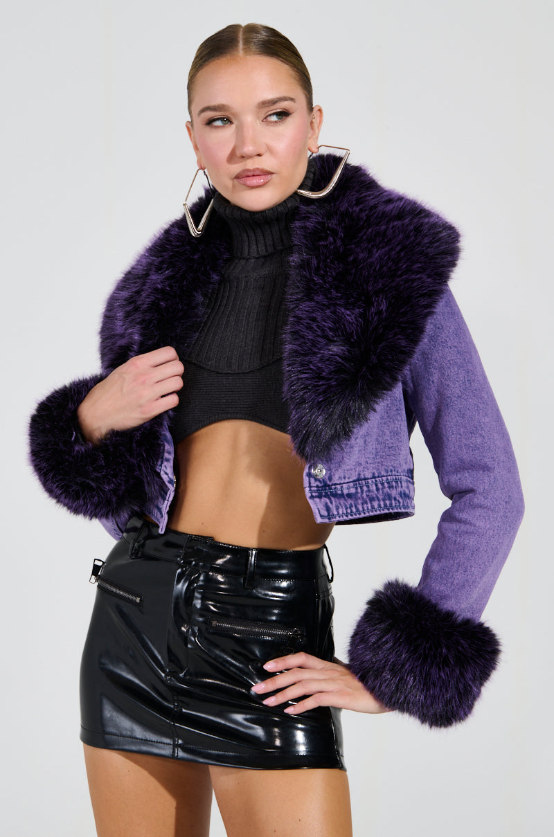 A person poses confidently wearing a Gisele Cropped Fur Trim Denim Jacket in purple over a black crop top and a shiny black miniskirt. They sport large hoop earrings, with their hair sleeked back, against a plain light gray background.