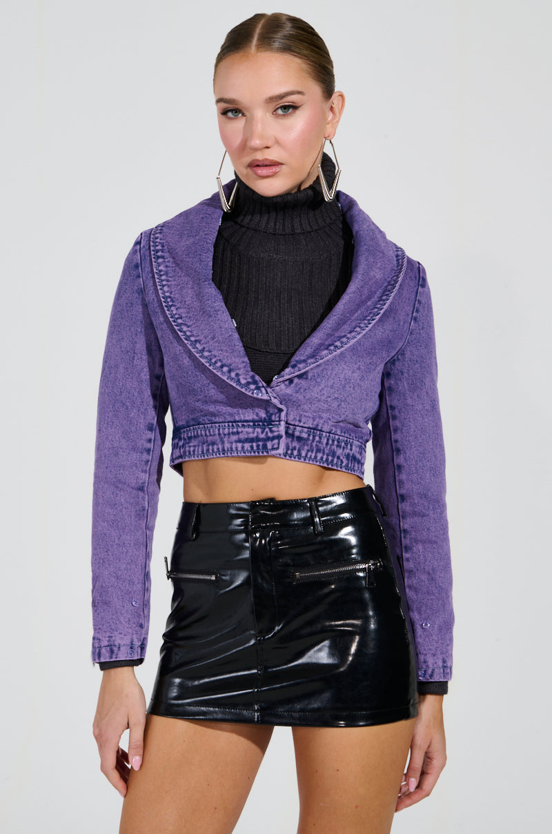 A person wearing the GISELE CROPPED FUR TRIM DENIM JACKET IN PURPLE, paired with a black turtleneck and a shiny black mini skirt, poses gracefully. The outfit is accessorized with large geometric earrings against a plain white background.