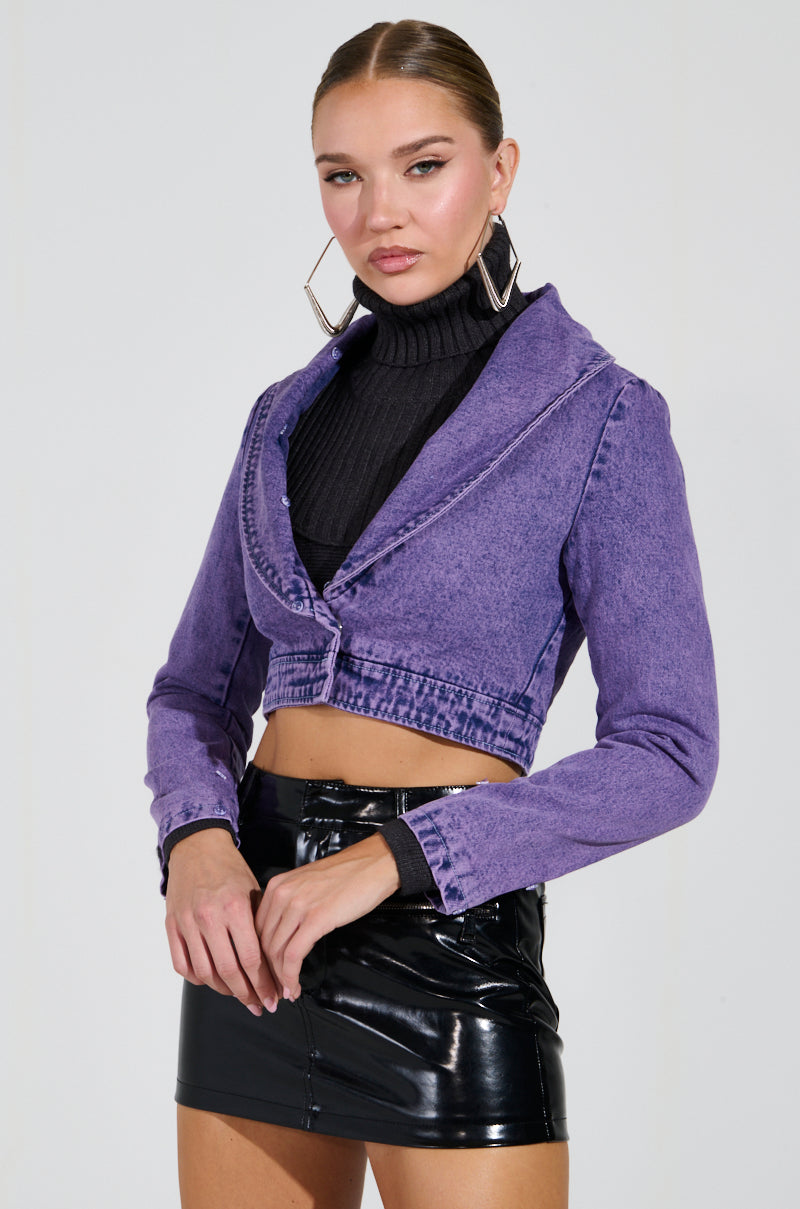 A person is styled in the Gisele Cropped Fur Trim Denim Jacket in purple, layered over a black turtleneck and teamed with a black leather mini skirt. They accessorize with large hoop earrings and wear their hair slicked back, standing against a simple background.