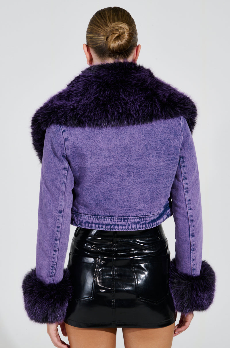 The person, with their hair tied back, is wearing the GISELE Cropped Fur Trim Denim Jacket in Purple, which features a plush faux fur collar and cuffs. They pair this stylish jacket with a shiny black leather skirt while facing away from the camera against a plain background.