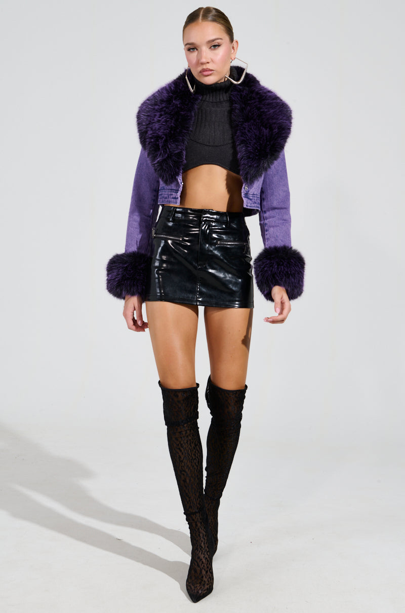 A person poses confidently in the GISELE Cropped Fur Trim Denim Jacket in Purple, paired with a black crop top, a glossy black mini skirt, and thigh-high patterned boots against a plain background.