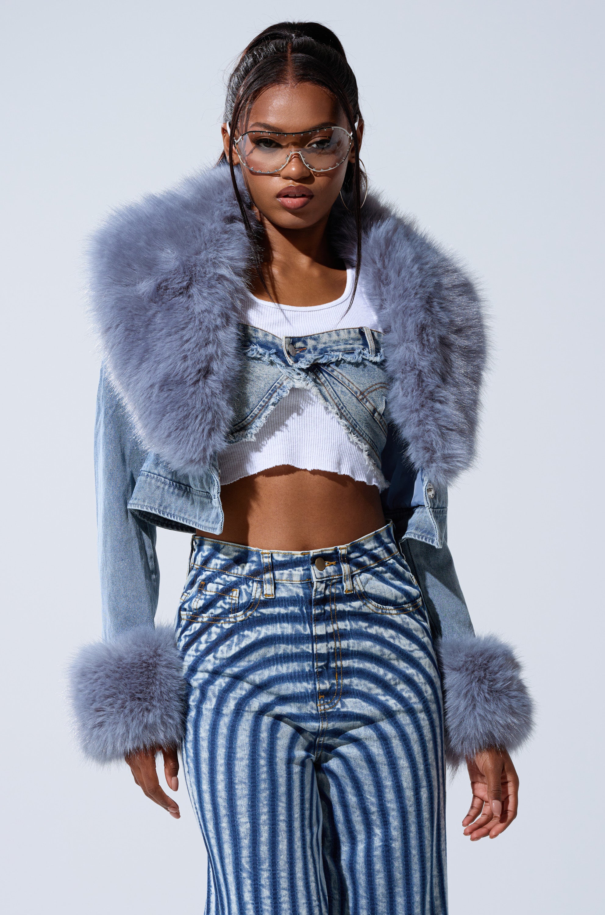 A woman channels Bratz Doll style in the GISELE CROP DENIM BLAZER with oversized light blue faux fur collar and cuffs, paired with a white crop top and high-waisted, blue-and-white striped denim pants against a plain background.