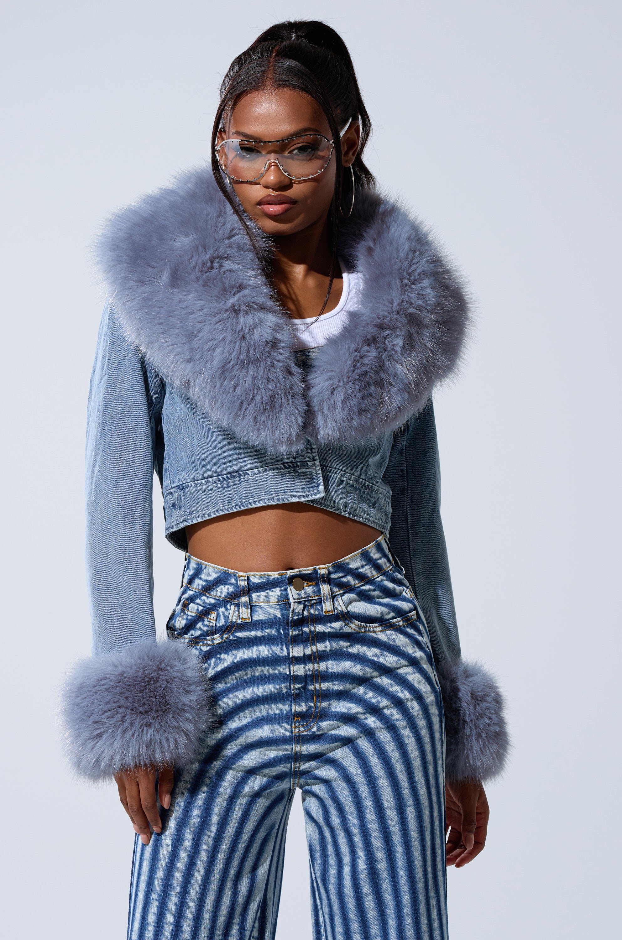 A woman wearing the GISELE CROP DENIM BLAZER with large, fluffy gray fur on the collar and cuffs, high-waisted blue-and-white striped pants, and clear glasses stands against a plain background.