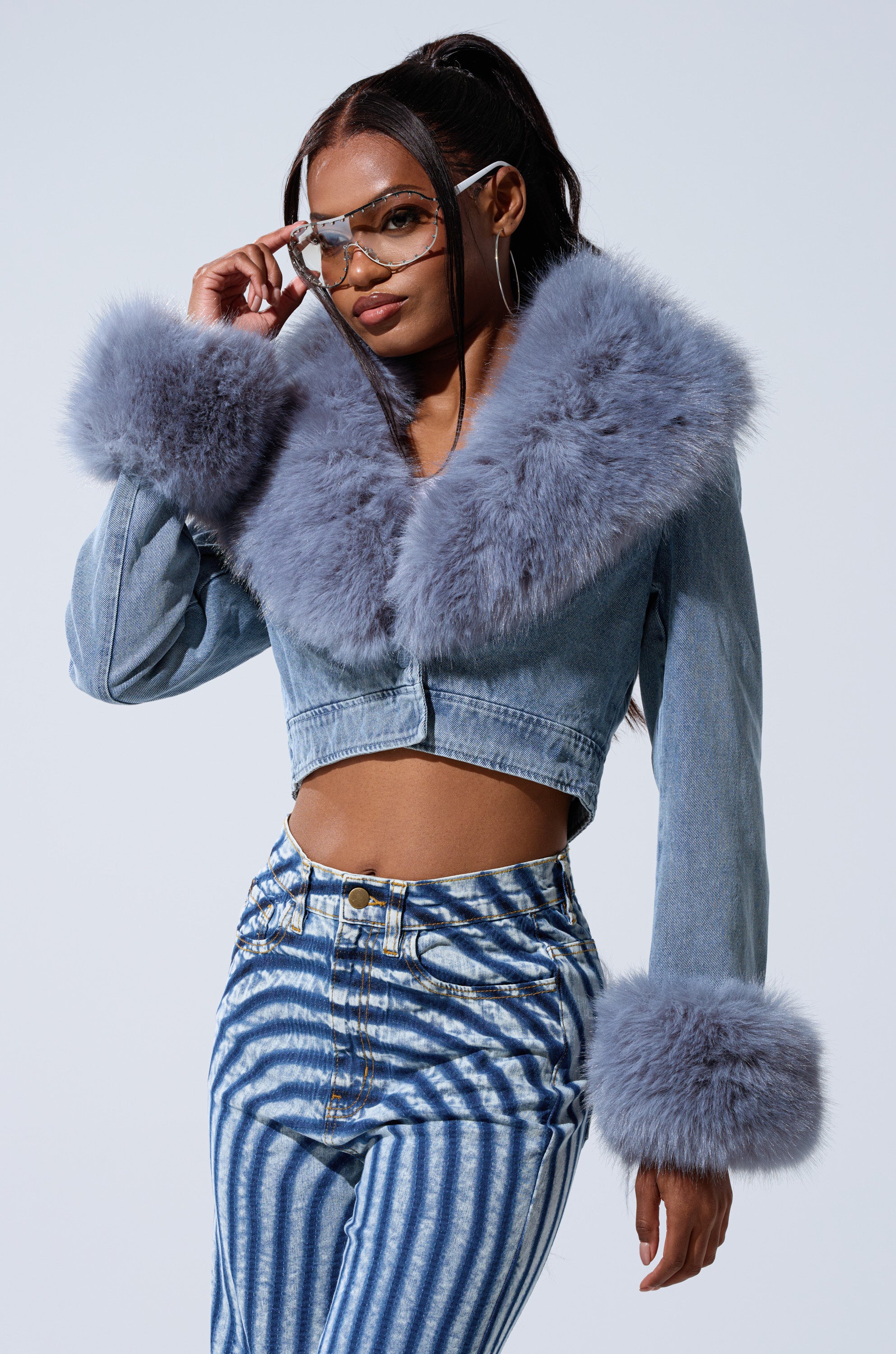 A woman in a Bratz Doll-inspired look wears the GISELE CROP DENIM BLAZER with large, fluffy gray fur trim, high-waisted striped jeans, hoop earrings, and tinted sunglasses as she poses confidently against a plain background.