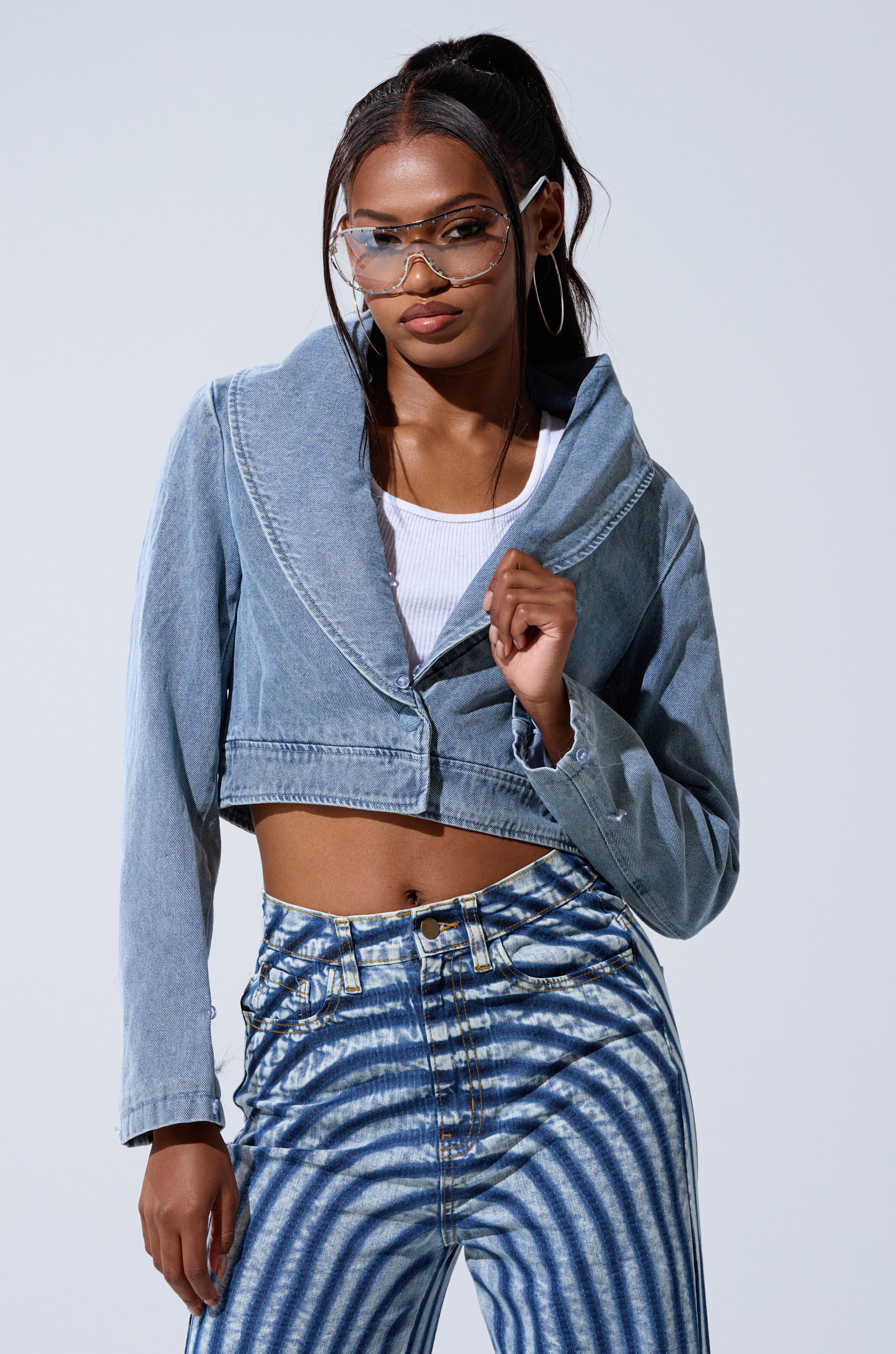A woman wears the GISELE CROP DENIM BLAZER over a white ribbed crop top and blue and white striped high-waisted jeans, accessorized with hoop earrings, clear sunglasses, and a high ponytail.