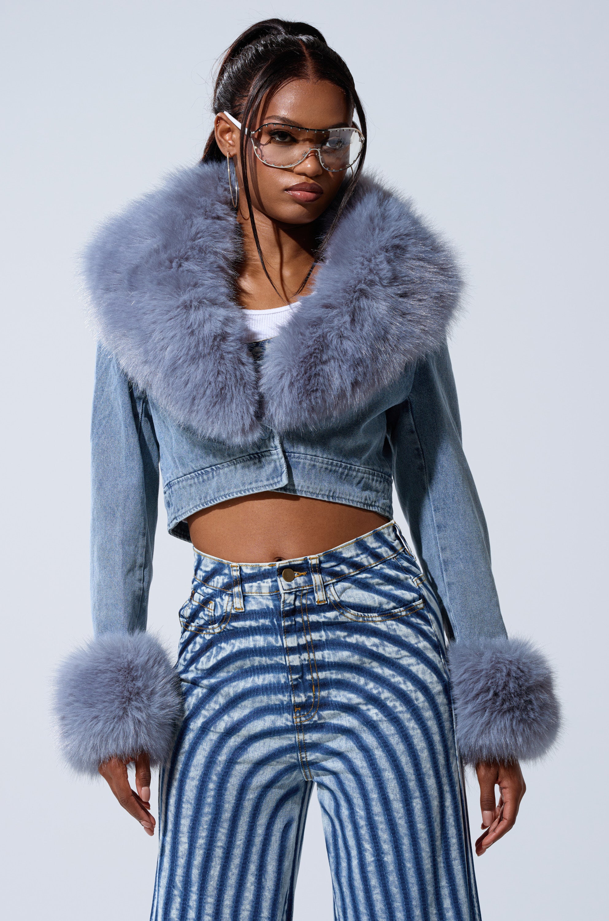 A person rocks Bratz Doll style in the GISELE CROP DENIM BLAZER—a light blue Y2K jacket with fluffy grey fur collar and cuffs—paired with large clear glasses, a white top, and high-waisted blue jeans with bold circular white patterns.